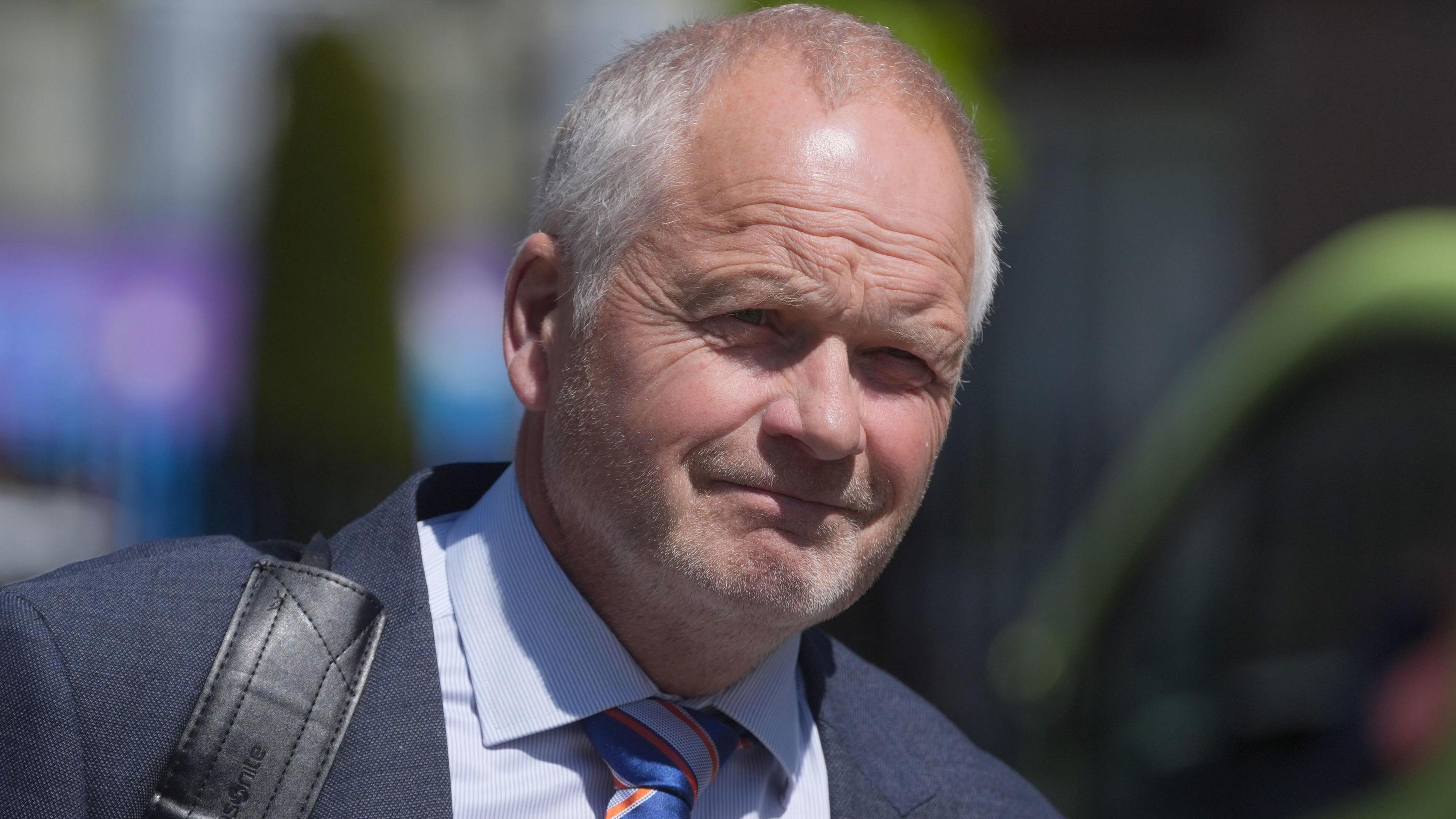Trevor Ringland is an older man with thin, short greying hair. He is squinting his eyes in the sun as he walks outside Dublin High Court. He is wearing a navy suit jacket, a striped white and light blue shirt and a striped blue and orange tie.