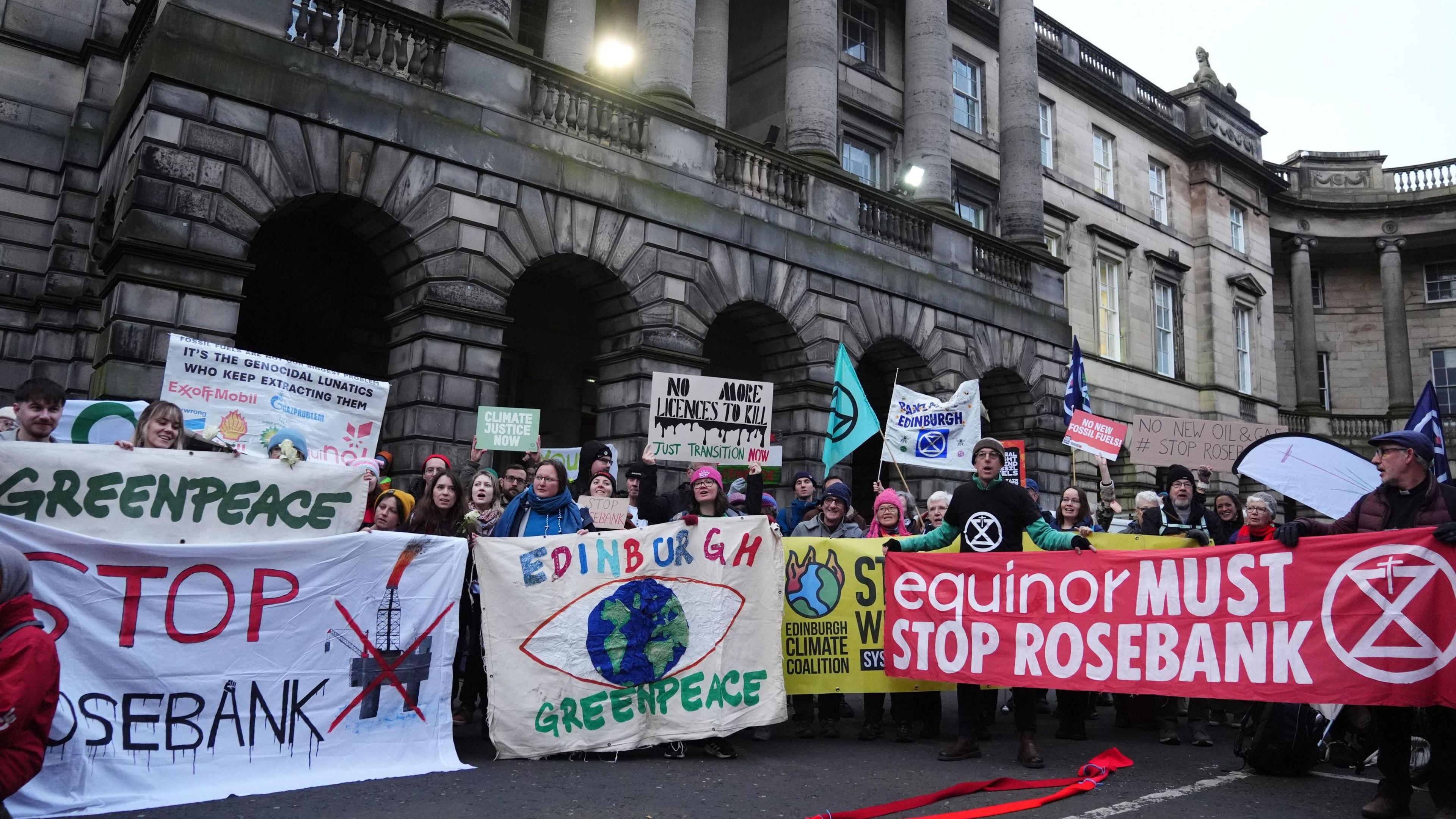 A group of protesters from climate organisations outside the Court of Session in Edinburgh. From left to right, one group are holding a greenpeace banner, which has the name of the organisation in green on a white banner. Below that is the words "Stop Rosebank" with stop in red and Rosebank in black. An image of an oil platform with a red X over the top is next to it. The next banner reads "Edinburgh" in multi-coloured letters and Greenpeace in green on a light-coloured banner either side of a globe in blue and green with a red eye shape surrounding it. The final banner is red with the words "equinor must stop Rosebank" in white letters.