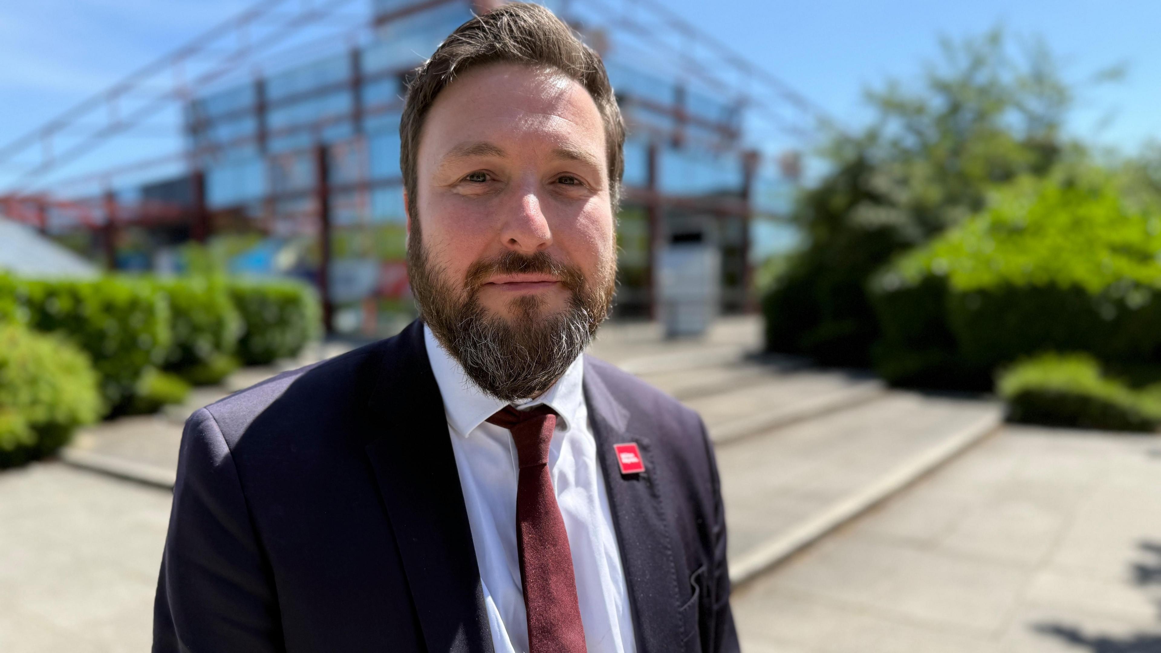 Pete Marland has a beard and dark hair, he is wearing a suit. Behind him is The Point.