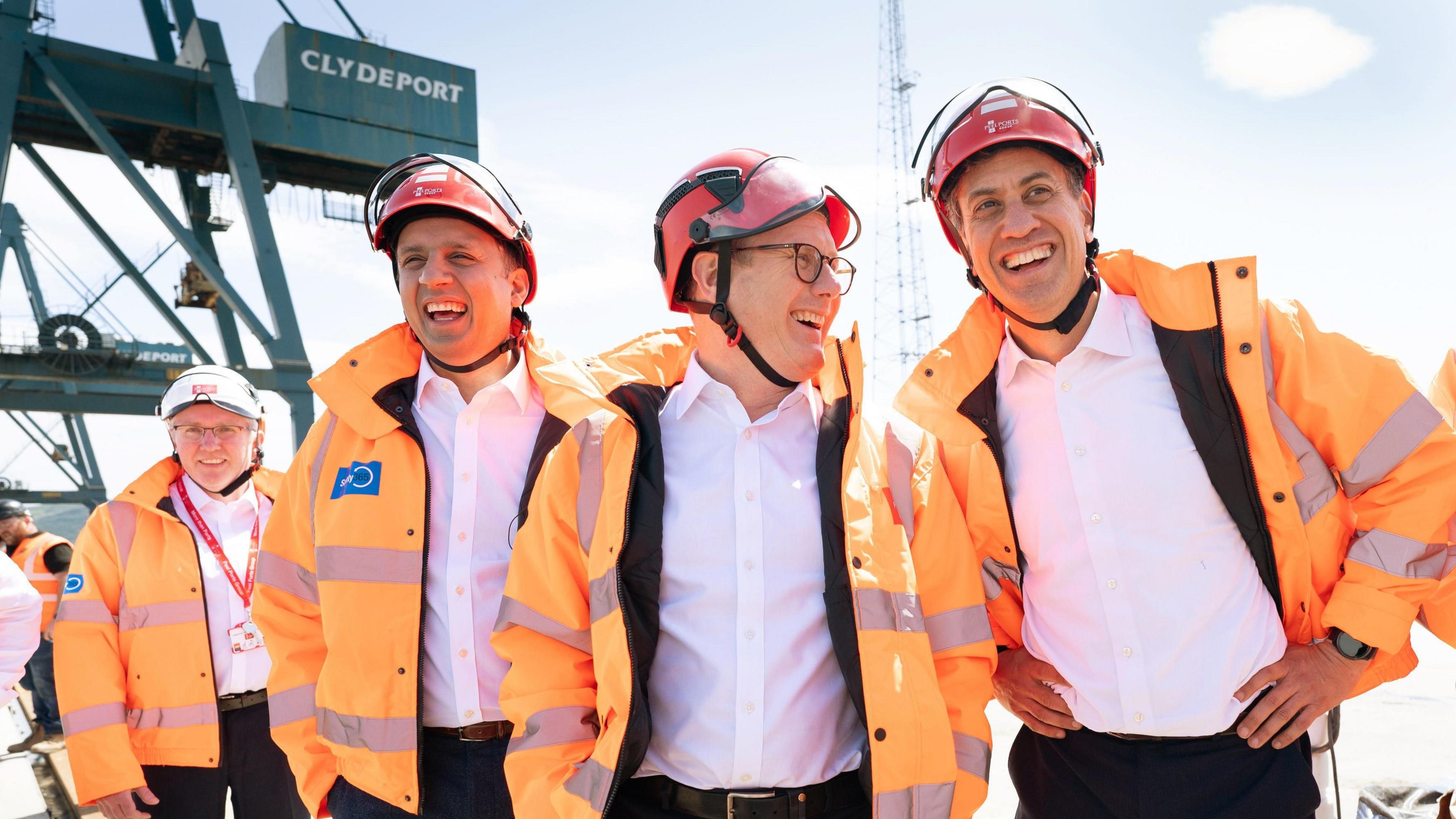 Anas Sarwar, Sir Keir Starmer, and Ed Miliband wear orange fluorescent jackets and red hardhats, as they stand in Clyde Port. Machinery is visible in the back-left.