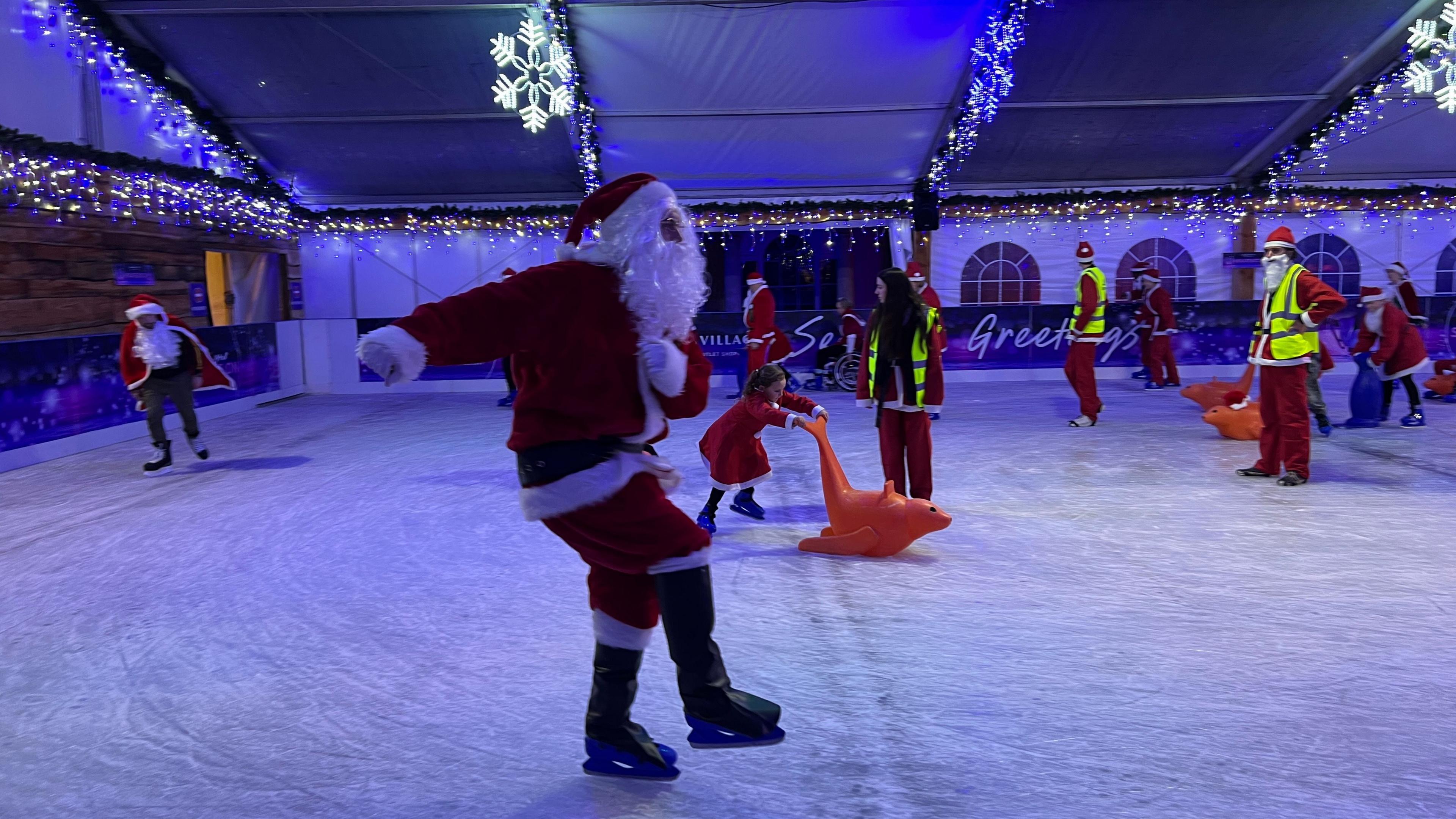 A person in a Santa costume is skating on an ice rink. There are more people in Santa costumes in the background.