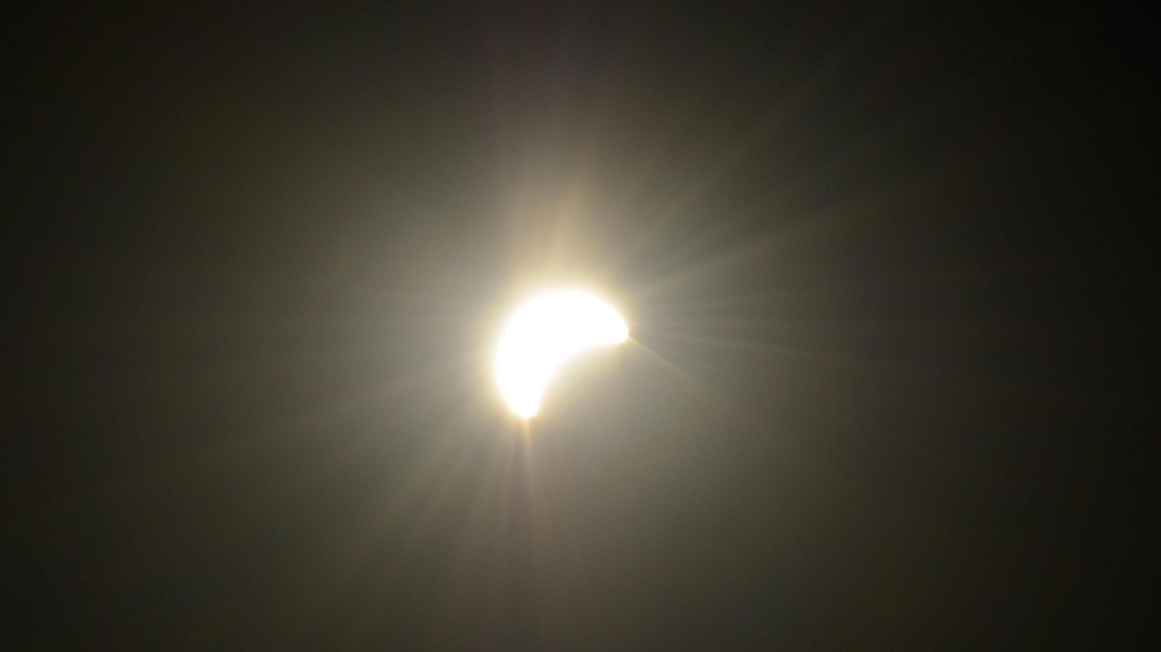 A close-up photograph shows the Sun during the eclipse as a bright crescent shape against a dark sky in Worthing, West Sussex. Rays of light extend from the Sun, while part of its disc is obscured by the Moon.