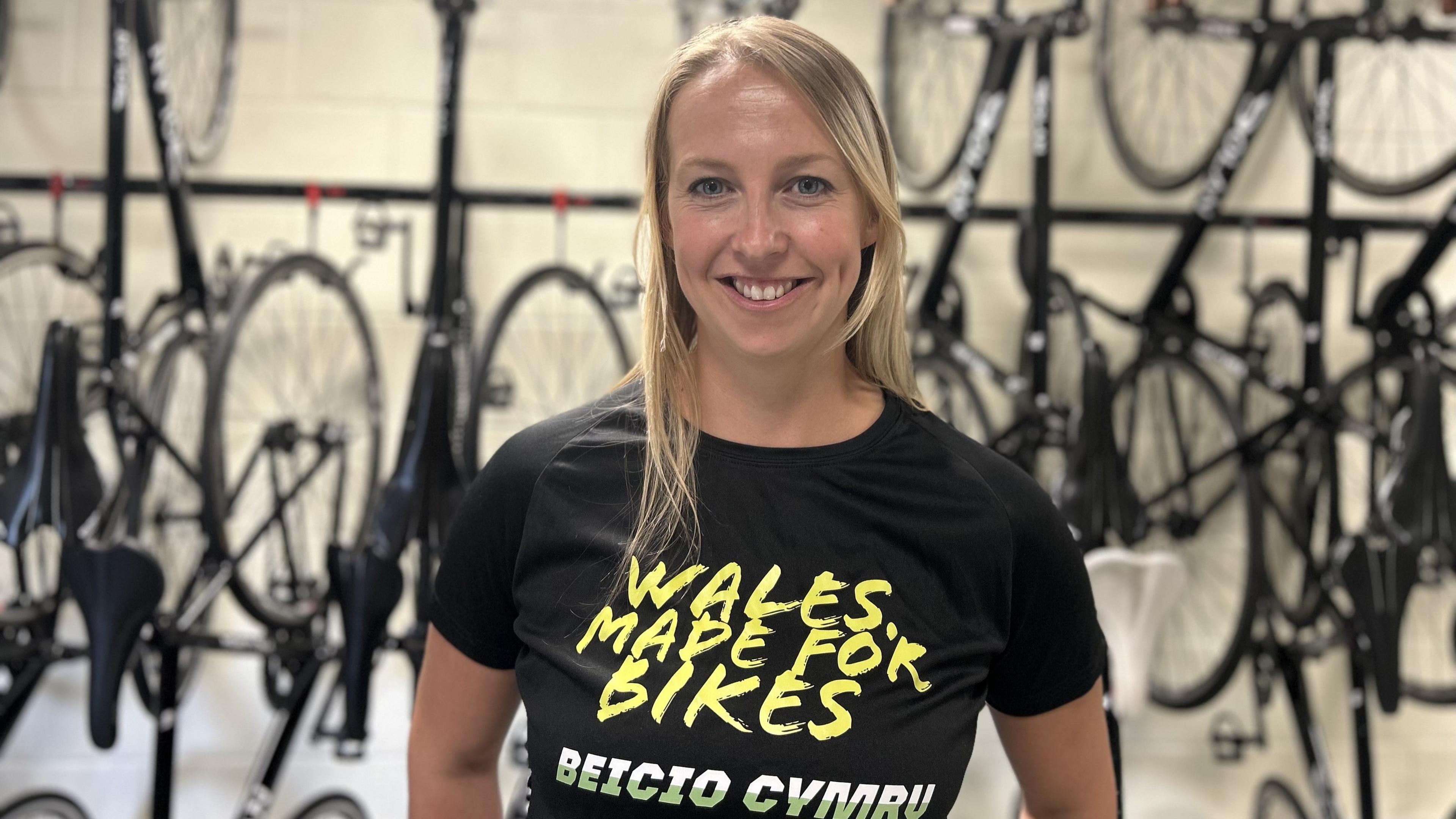 Rachel Draper yn edrych at y camera ac yn gwenu. Mae na feics ar y wal tu ol hi. Mae hi'n gwisgo crys-T du swyddogol Beicio Cymru sy'n dweud 'Wales. Made For Bikes'