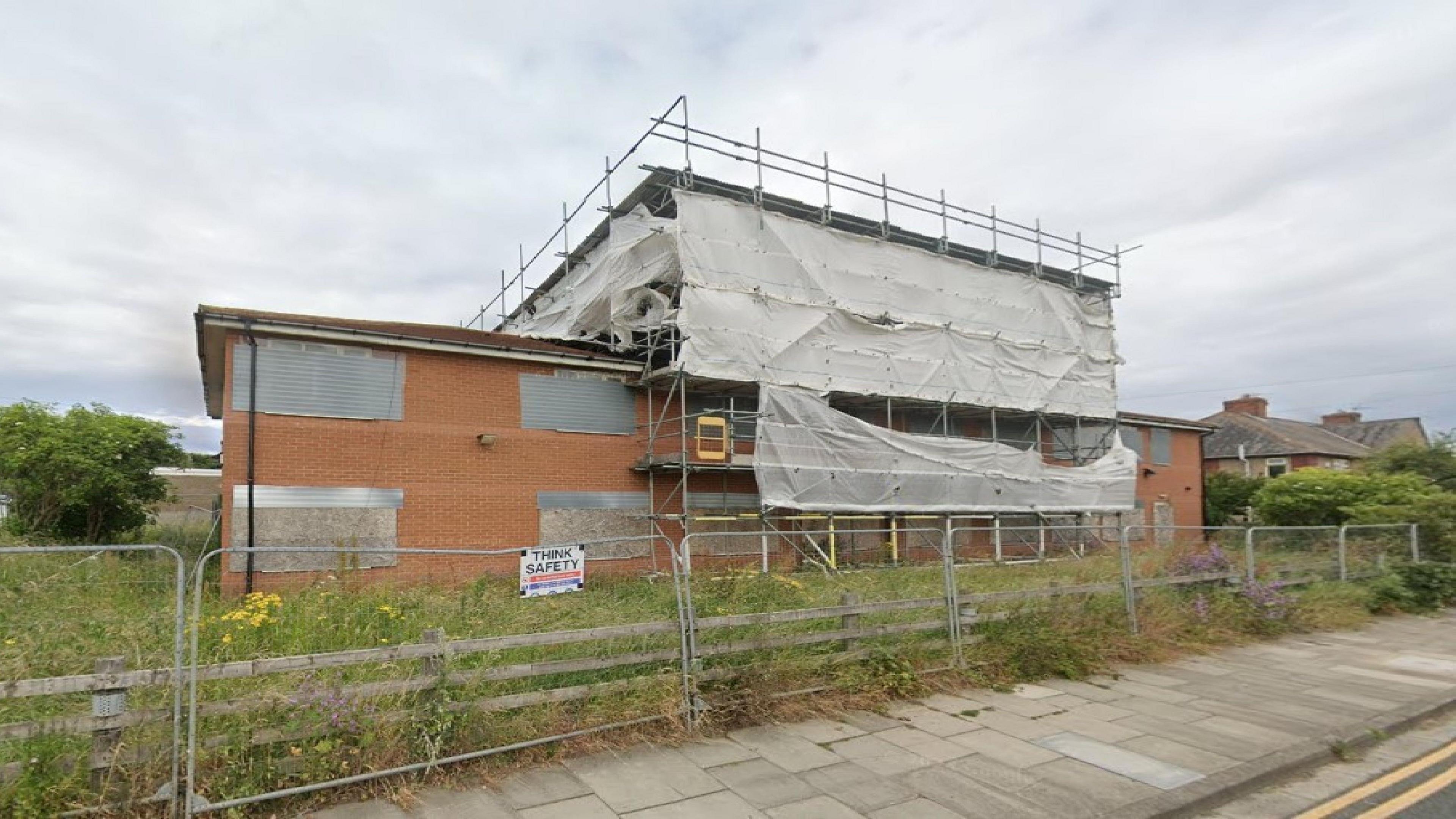 A derelict brick building, with windows that have been boarded up and scaffolding, stands in an overgrown field. 