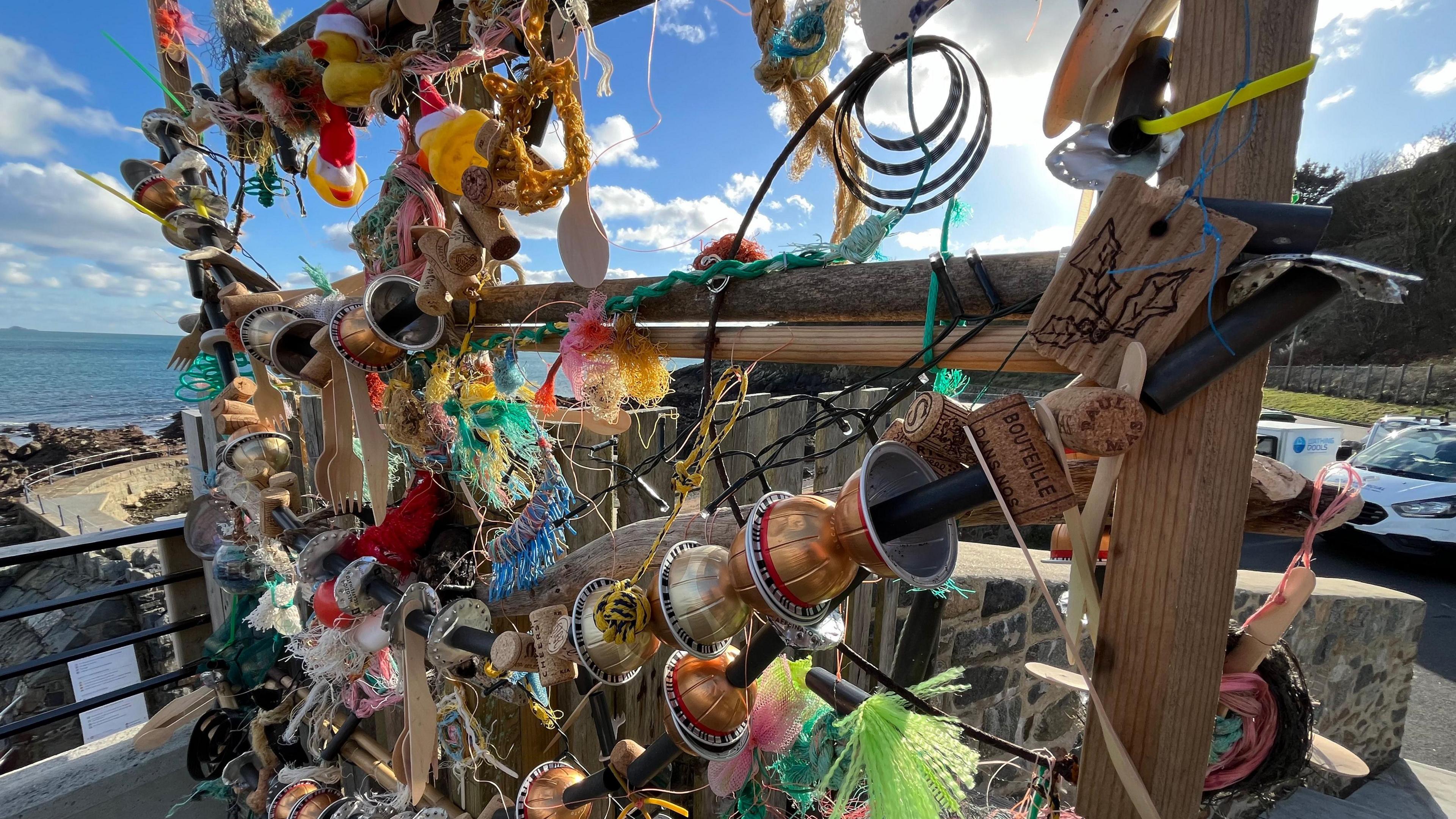 A close up of the Christmas tree with the sea in the background. Items on the tree include used coffee pods, disposable cutlery, corks, rubber ducks and fishing wire