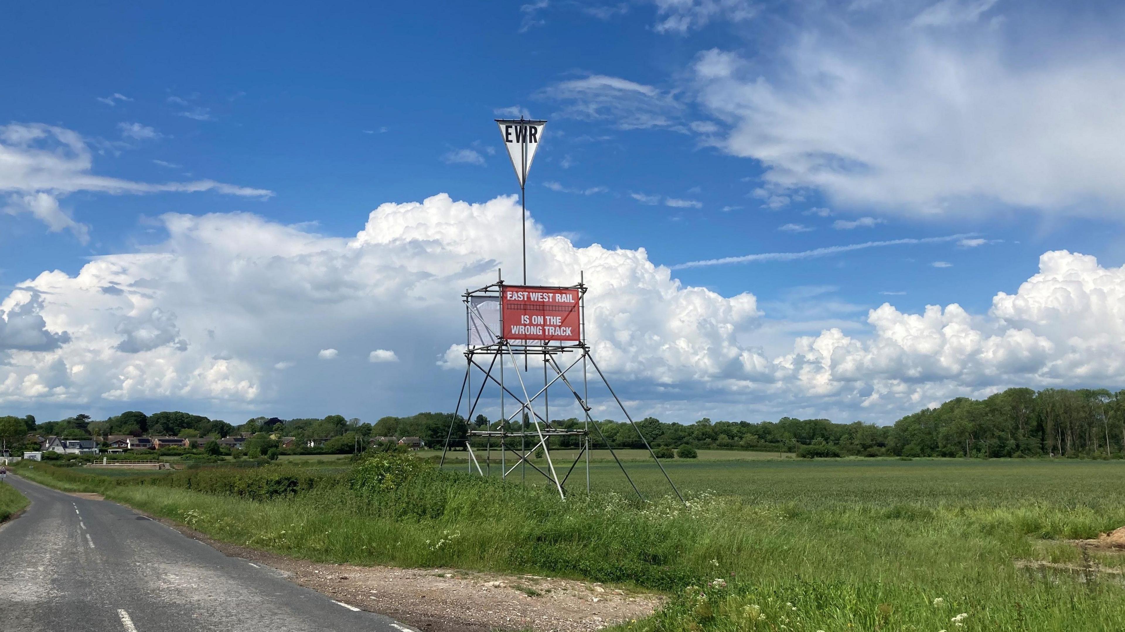 Sign on a pylon saying East West Rail is on the wrong track in a field beside the road