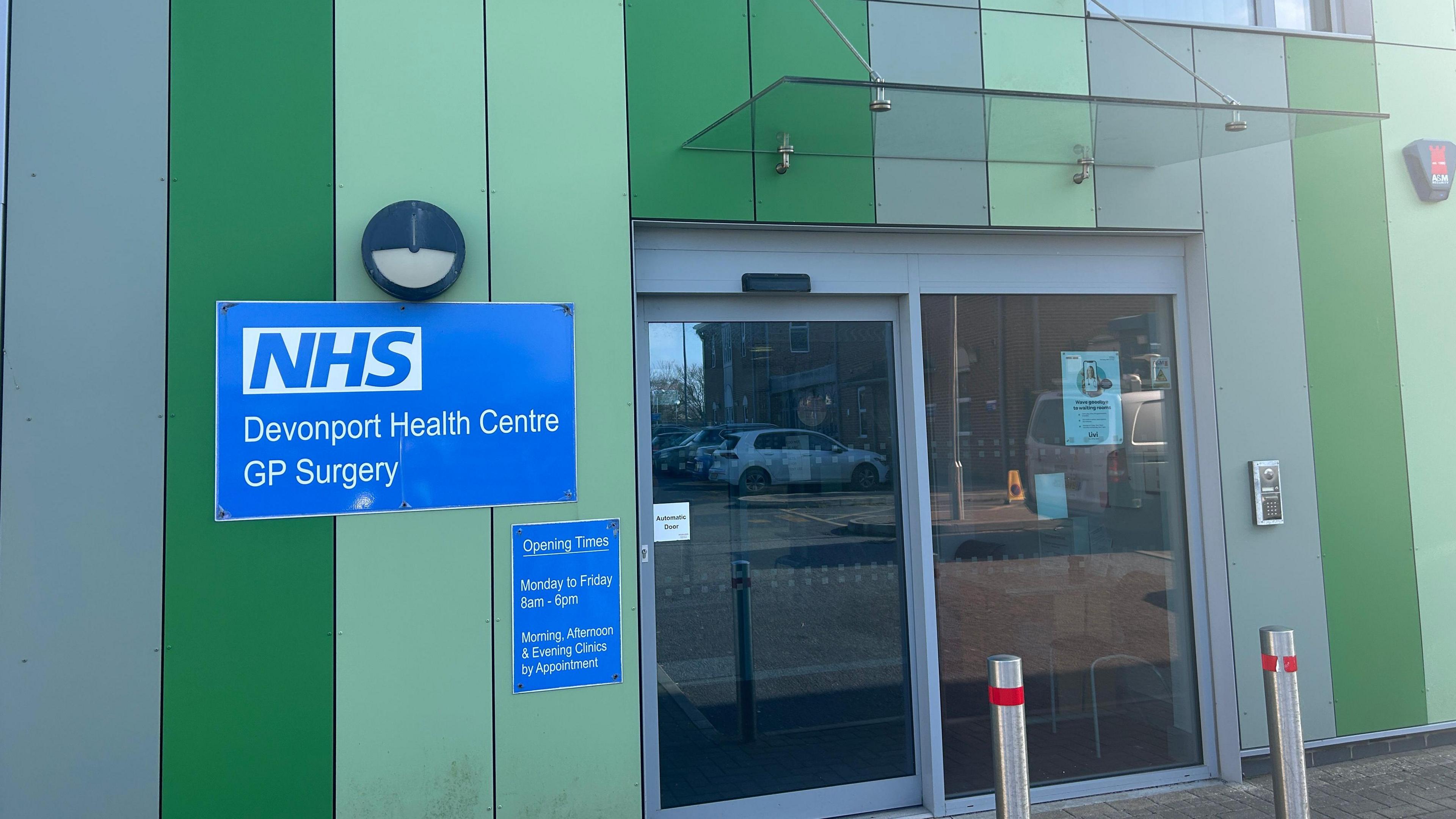 The front of a building which is modern and green. It has sliding automatic doors and a blue sign saying Devonport Health Centre GP Surgery 