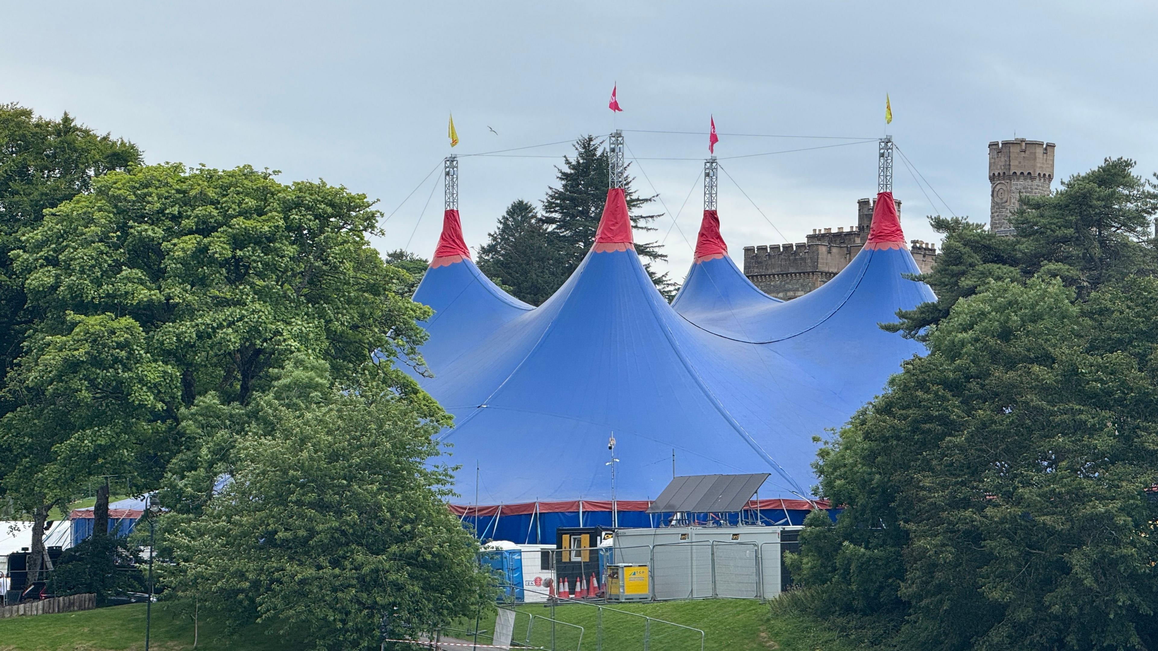 Dealbh a' sealltainn teanta mhòir ghuirm le pìosan beaga dearg air a' mhullach a tha air a dhol an-àirde sa Ghearradh Chruaidh ann an Steòrnabhagh airson Fèis Cheilteach Innse Gall. Tha craobhan mòra uaine timcheall orra agus pàirt den Chaisteal air a chùl. 