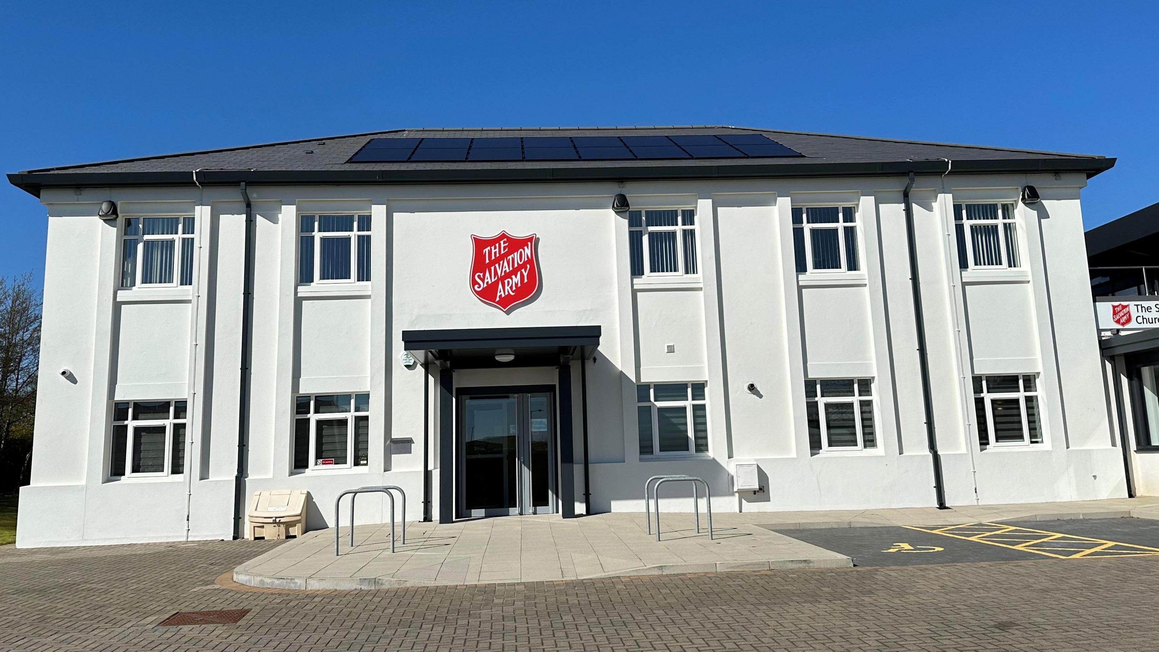 A large white building with a glass door entrance that has a red Salvation Army badge above it. There is paving in front of the building along with disabled parking spaces marked out in yellow paint.