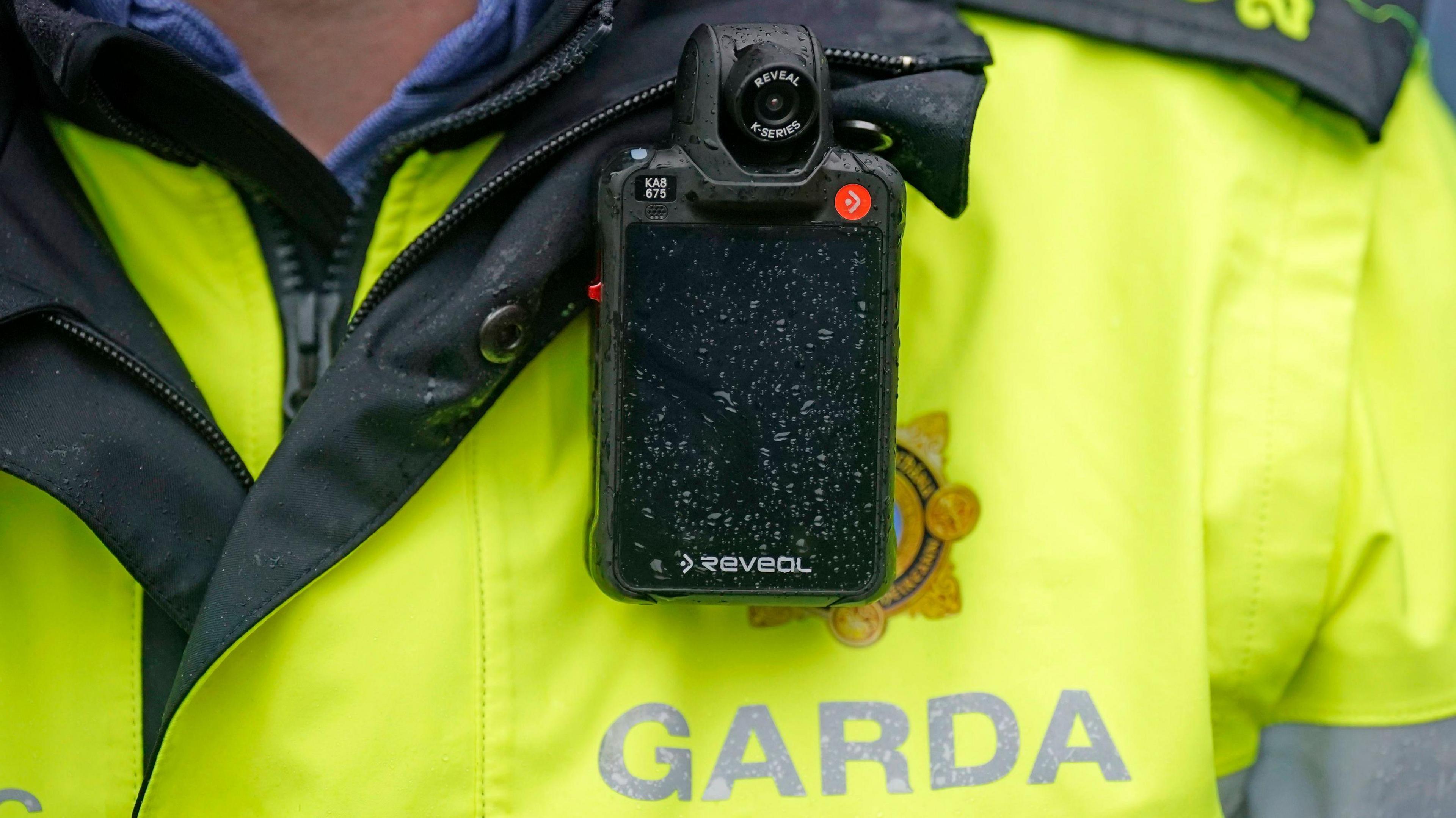 A close up of a Garda uniform. Garda is written in grey letters on a bright yellow jacket. A camera is attached to the uniform. 