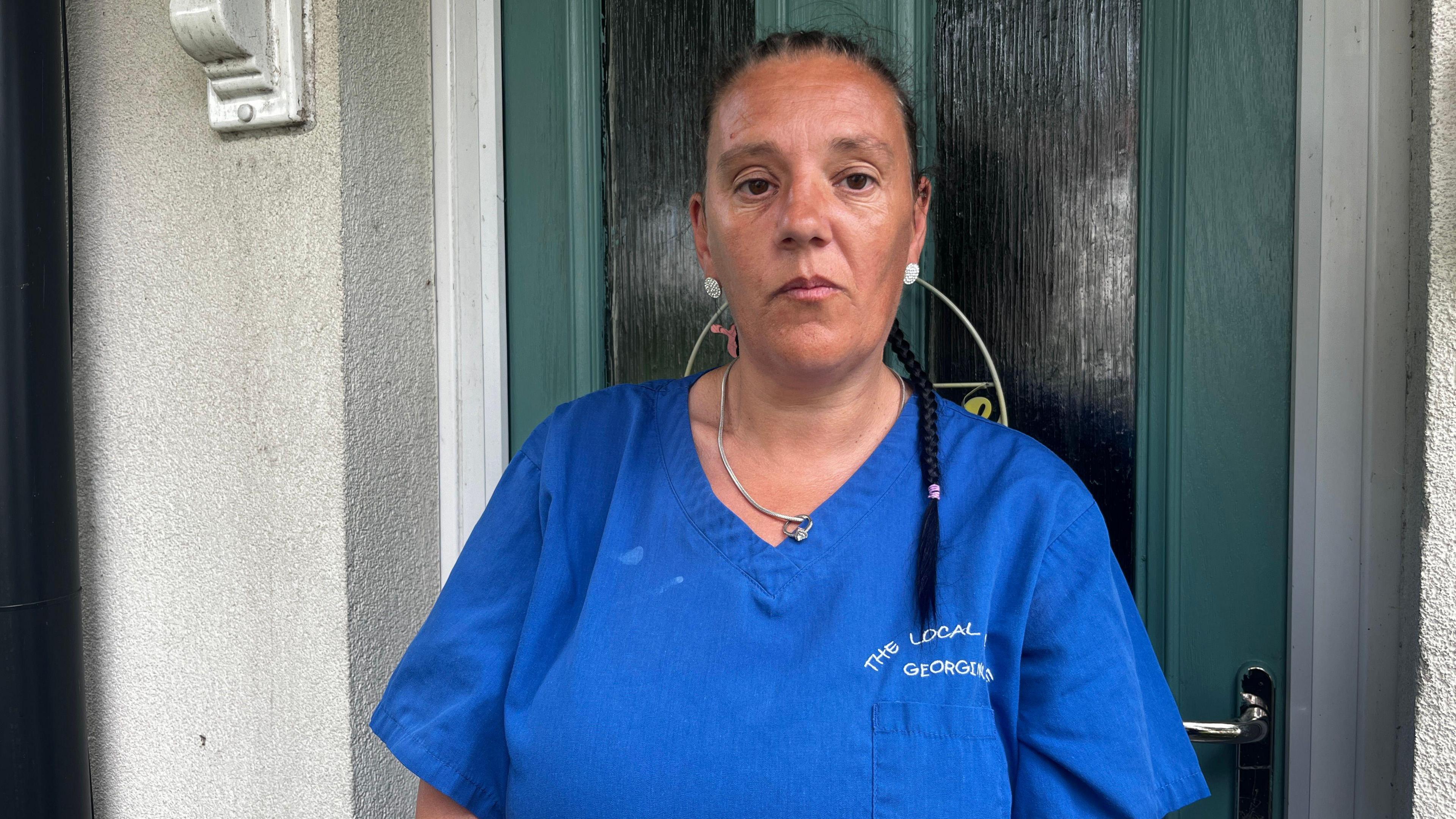Georgina Thomas, 39, who has black hair tied into a braid and is wearing blue scrubs, stands in front of a green front door.