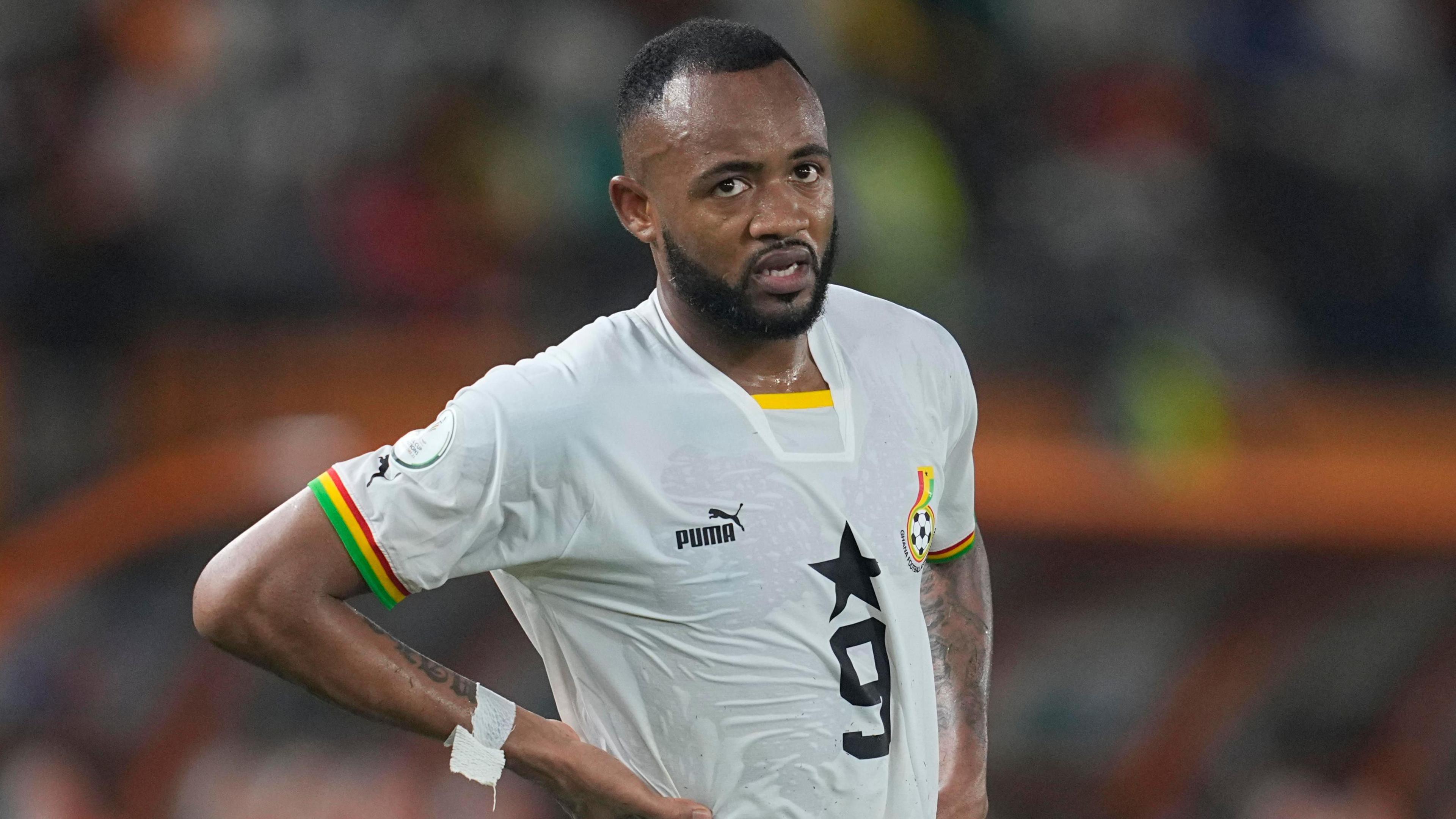 Jordan Ayew, wearing a white Ghana shirt with a number nine on the chest, stands with his hands on his hips