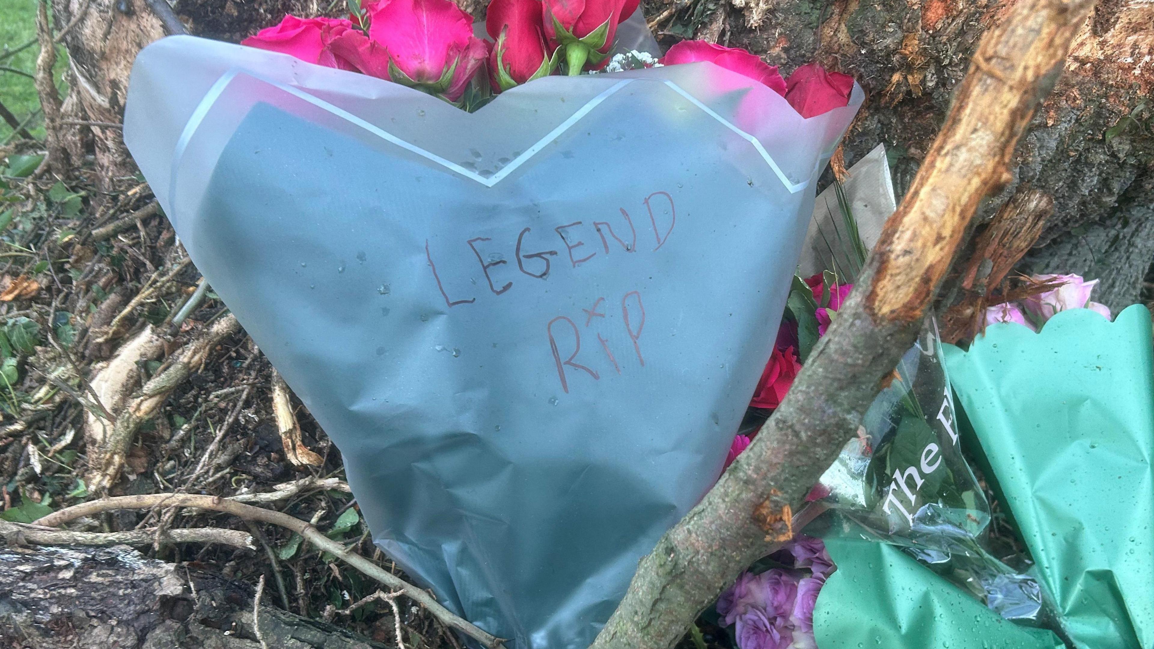A close up of bouquets of flowers lying beside a tree. Legend RIP is written on the cover of a bouquet of pink roses. There is an x above the i in RIP. 