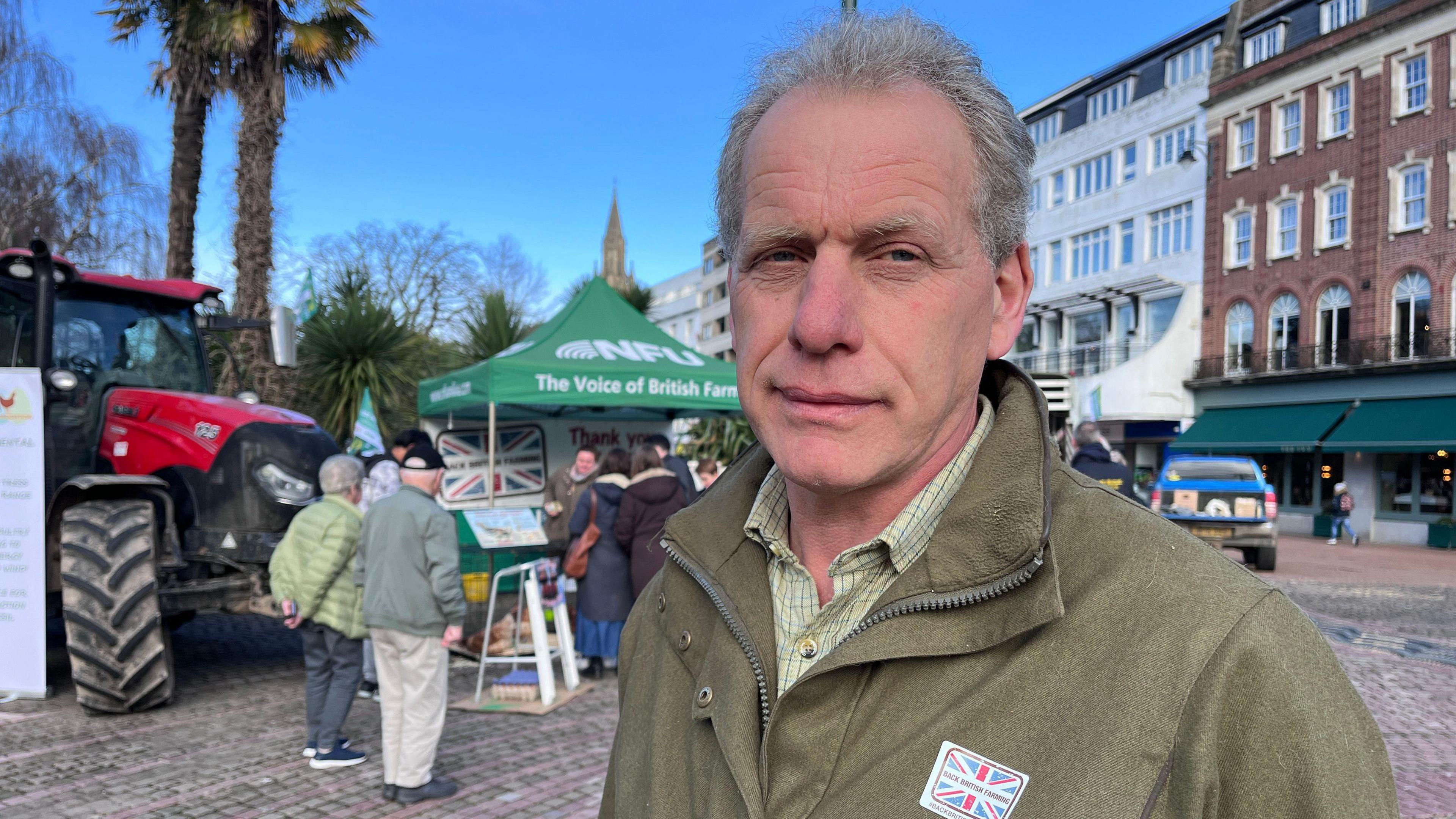 Tim Gelfs wearing a khaki jacket stood in front of a green NFU stall and a red tractor