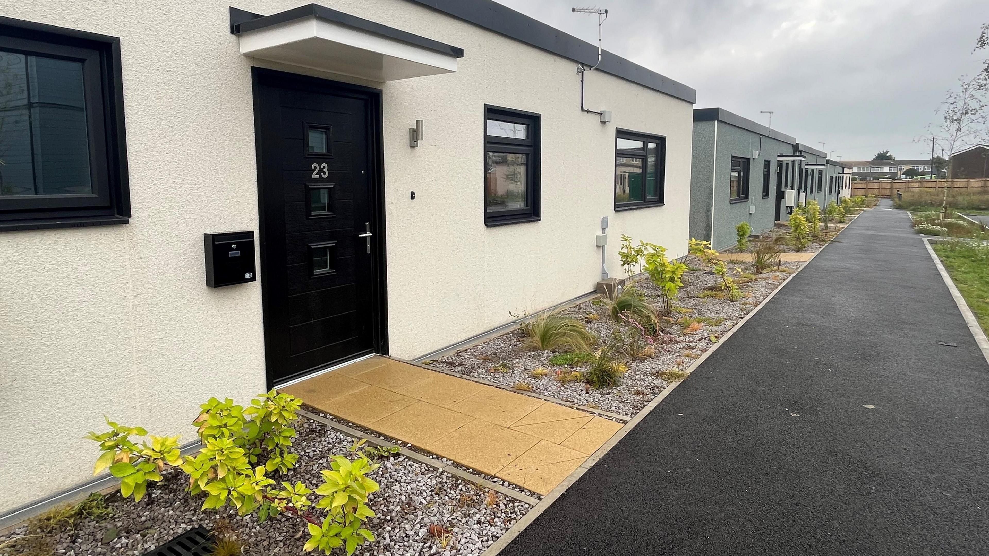 The front entrance of a portable cabin with a paved walkway and landscaped borders with shrubs and gravel