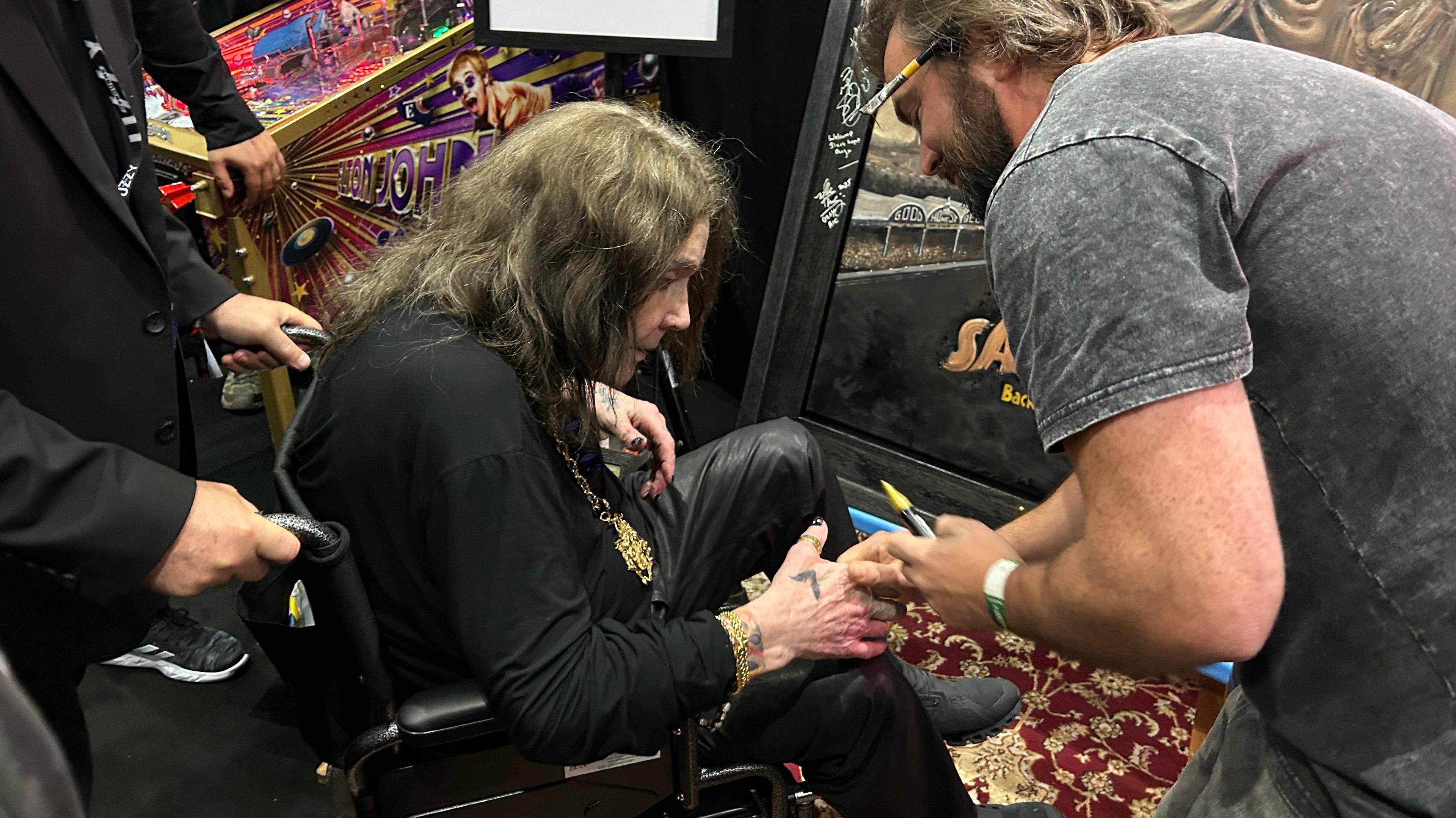 Ozzy Osbourne, in a wheelchair, is shaking hands with a man who is stood up. The man is holding a yellow marker pen.