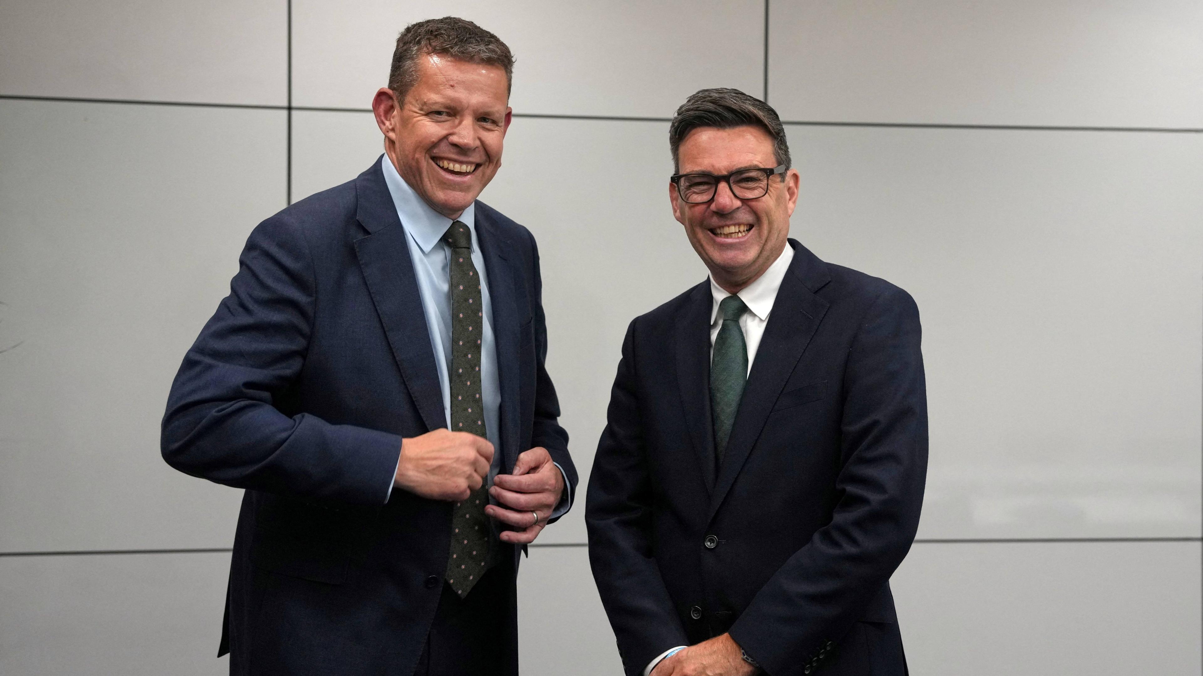 Rhun ap Iorwerth, a tall man with fair hair, is smiling broady whole buttoning the jacket on his dark blue suit. He is wearing a light blue shirt and green patterned tie. Beside him Andy Burnham is in a dark blue suit with white shirt and solid green tie and is wearing black plastic-rimmed spectacles. They are both laughing.