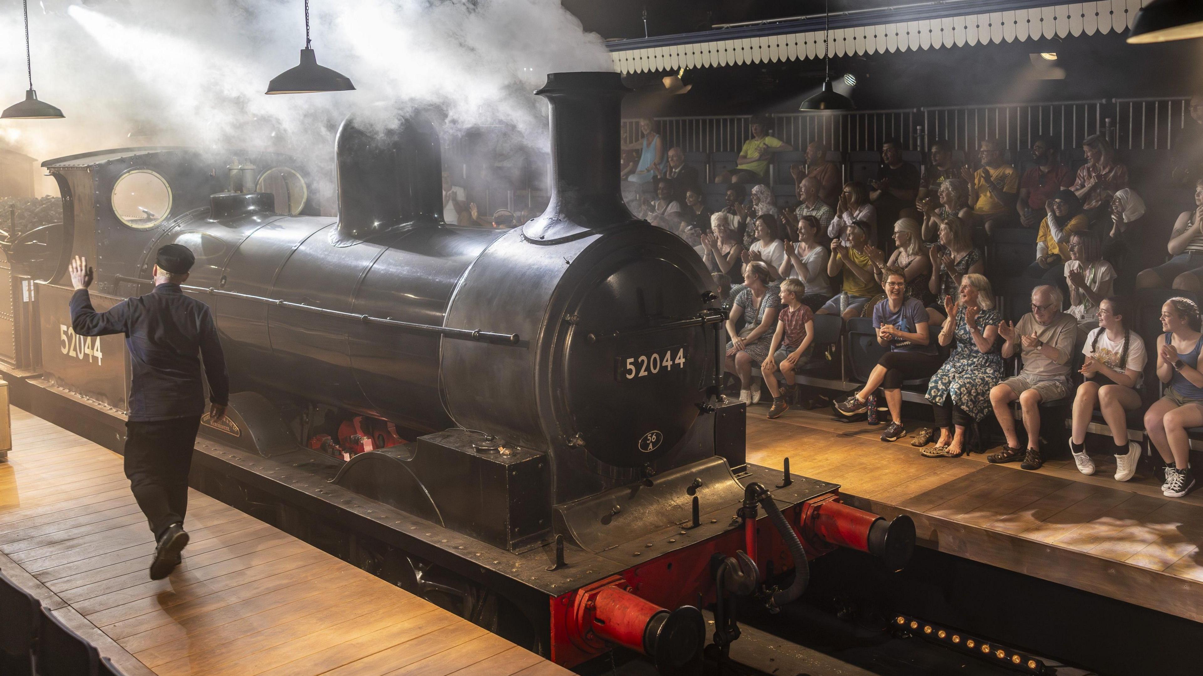 A picture of a black steam train with the number 52044 on the front and side the audience seated behind it in a specially adapted theatre built in an engine shed on the Keighley and Worth Valley
