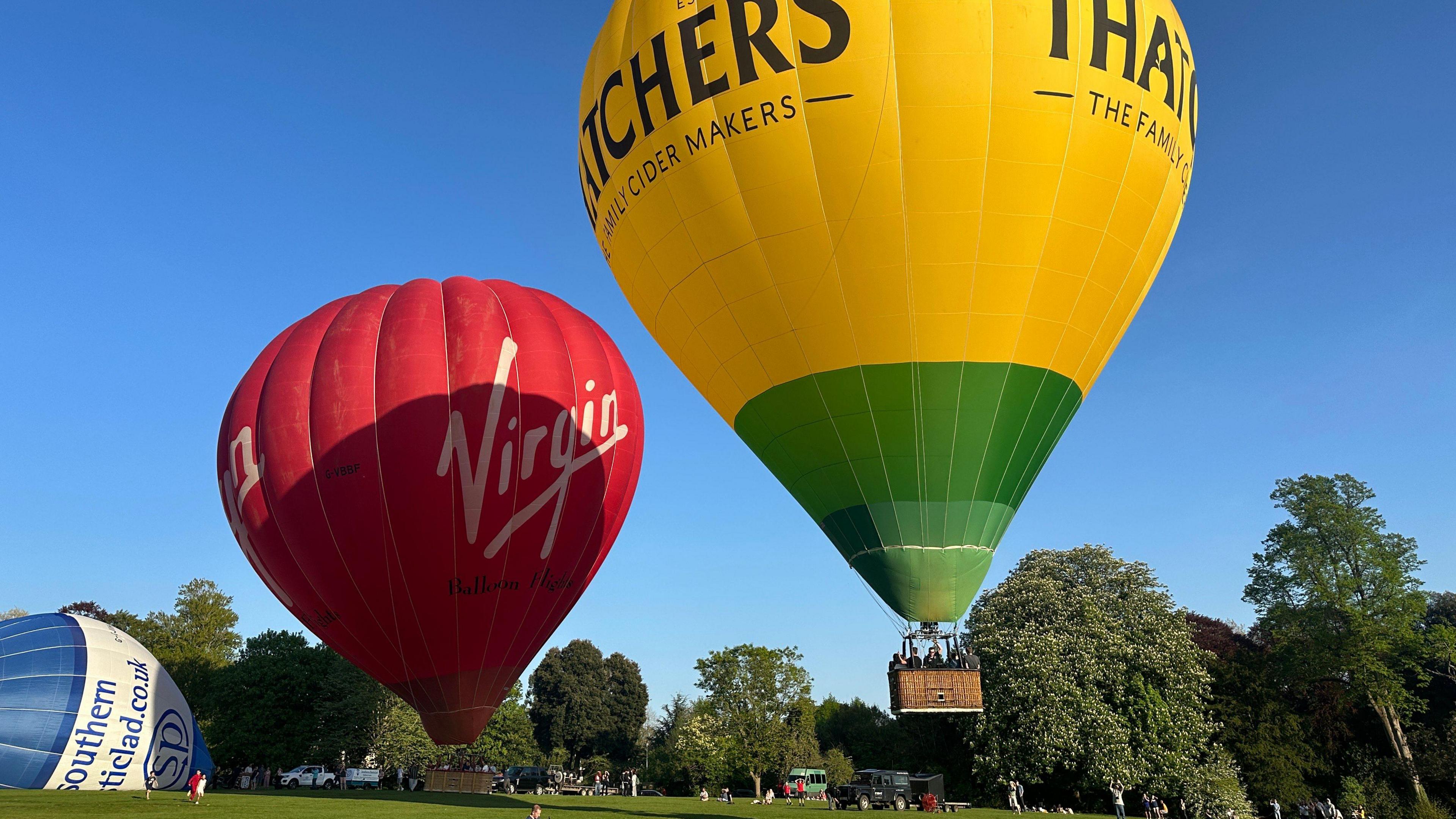 A Virgin-branded hot air balloon and a Thatchers -branded balloon hover over a playing field dotted with trees. A partially inflated blue and white balloon is visible to the left of the shot.  