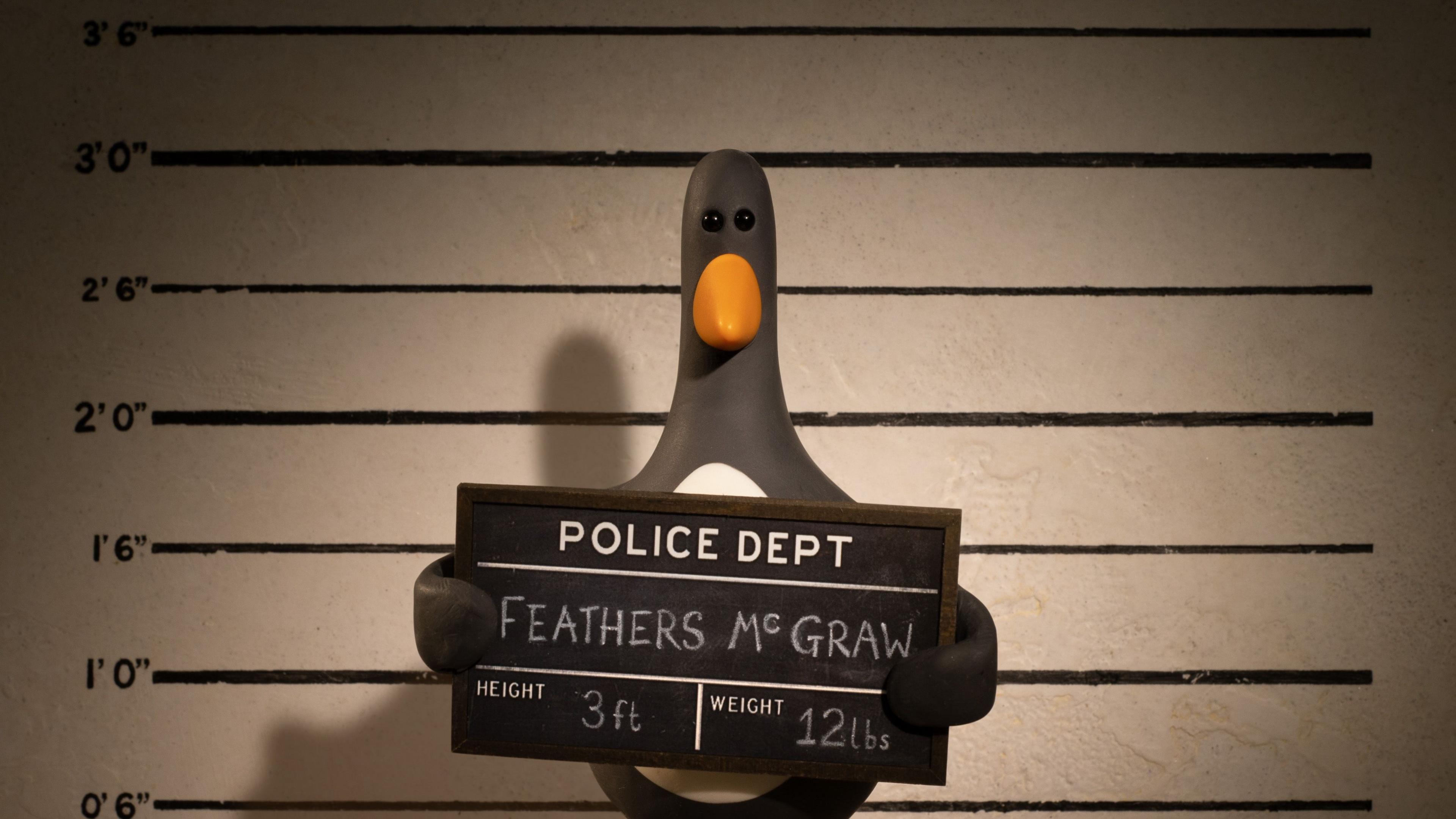 A model of Wallace and Gromit character Feathers McGraw. The penguin character is holding up a chalk board with his name, height and weight on and standing in front of a police suspect board. 