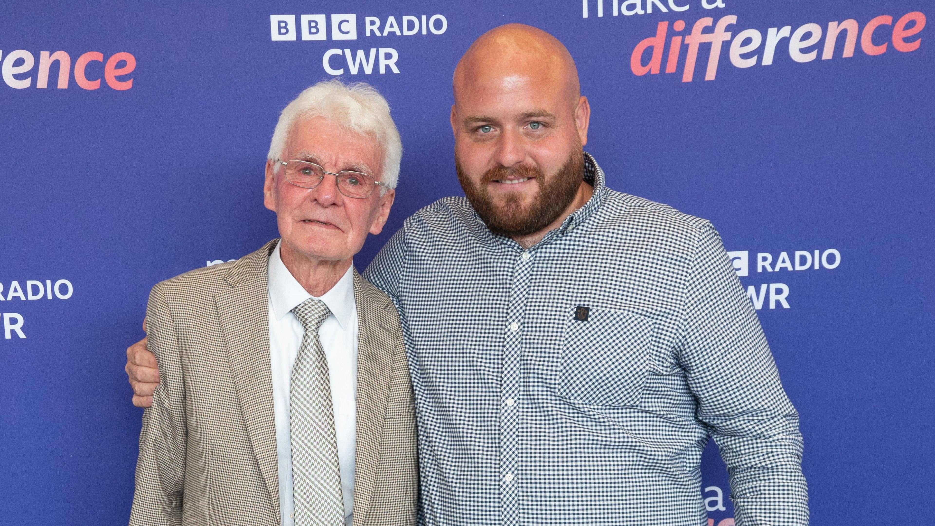 Steve is standing on the right wearing a blue and white shirt. He has a bald head. He has his arm around a man with white hair and glasses, who is wearing a suit