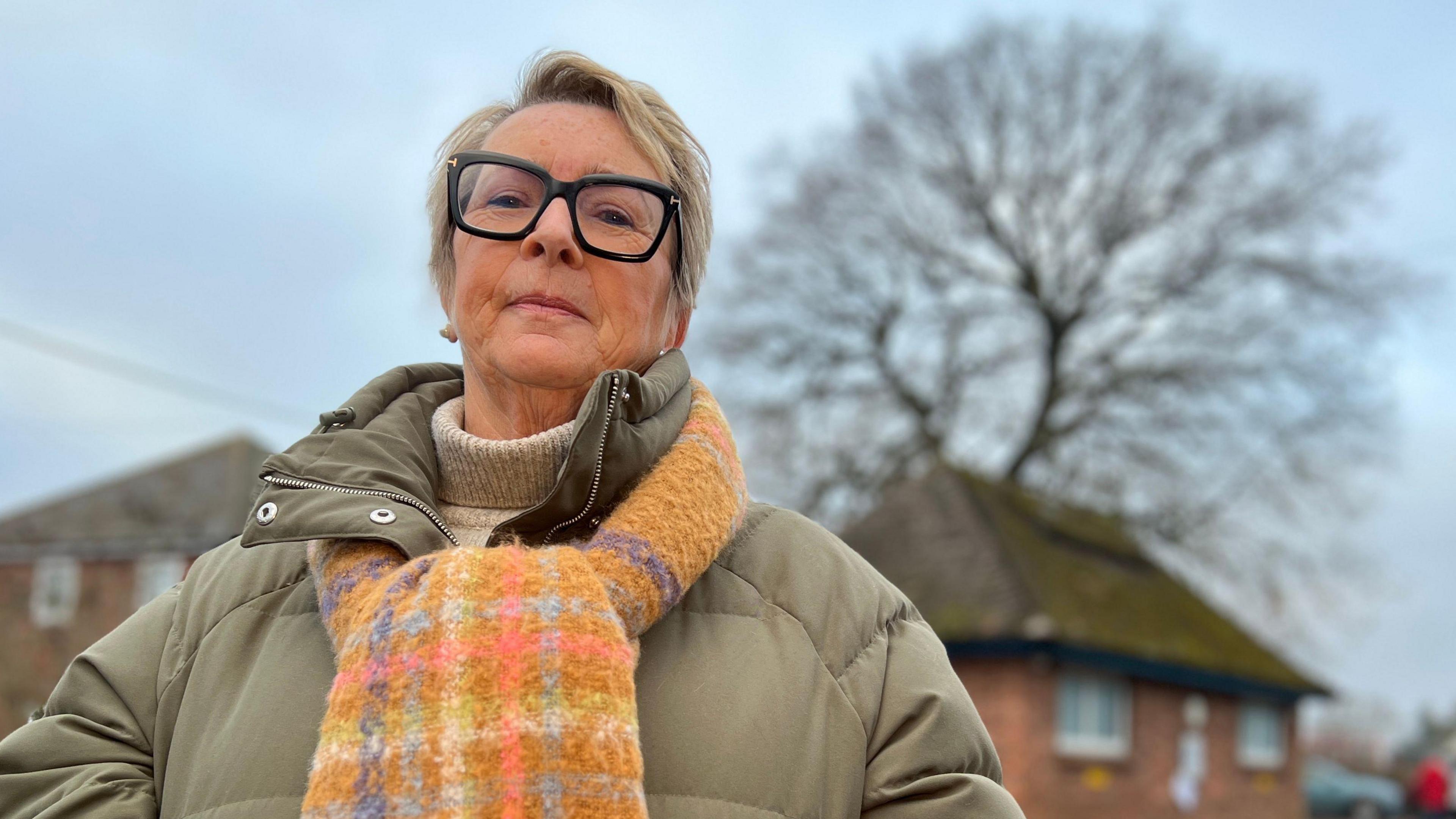 Councillor Denise Burke stood in a car park with a large oak tree in the background. She is wearing a green coat with an orange scarf, and has dark rimmed spectacles on her face.