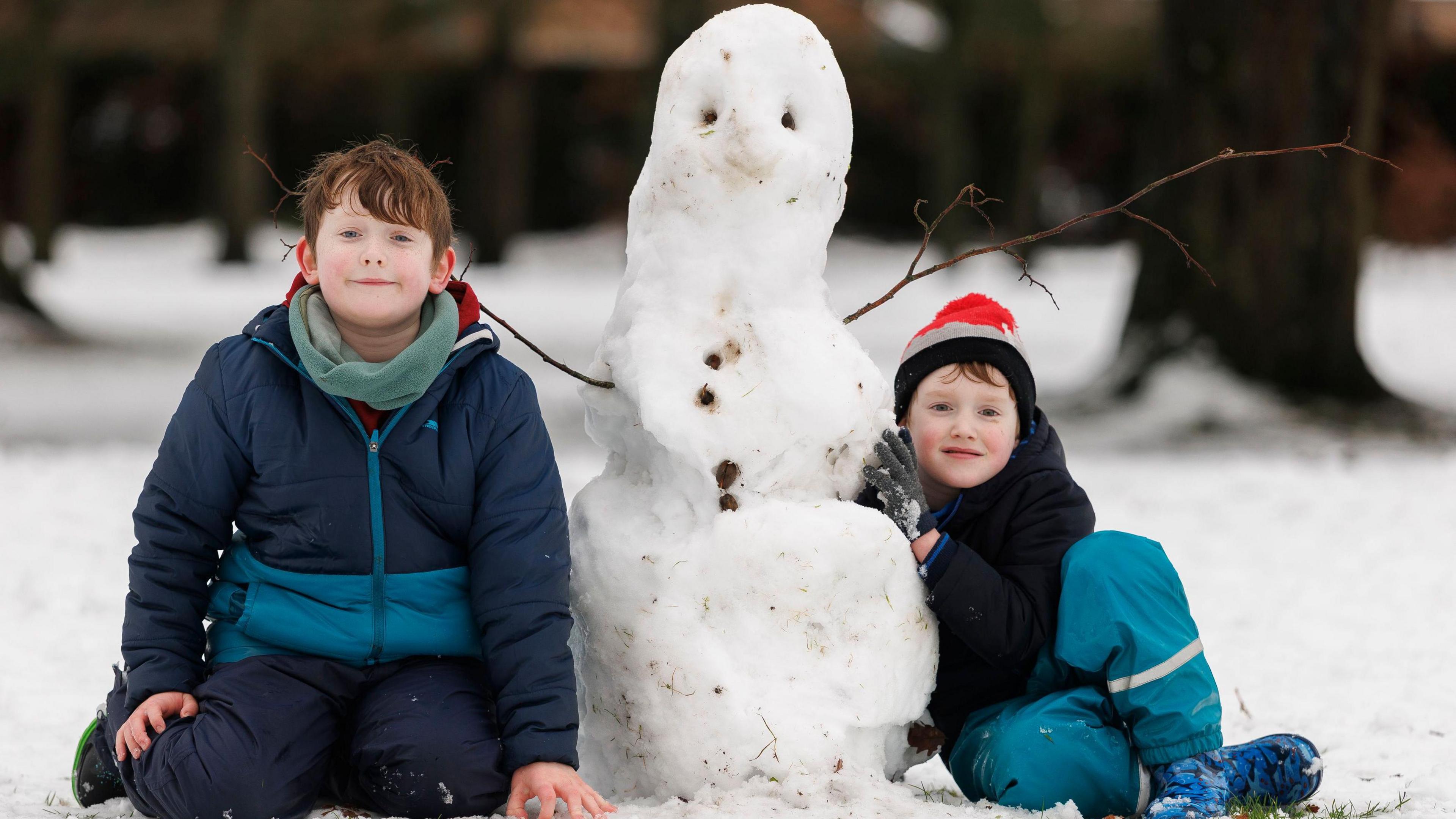 Two young boys dressed in warm weather gear are sitting on snowy ground on either side of a snowman they have built 