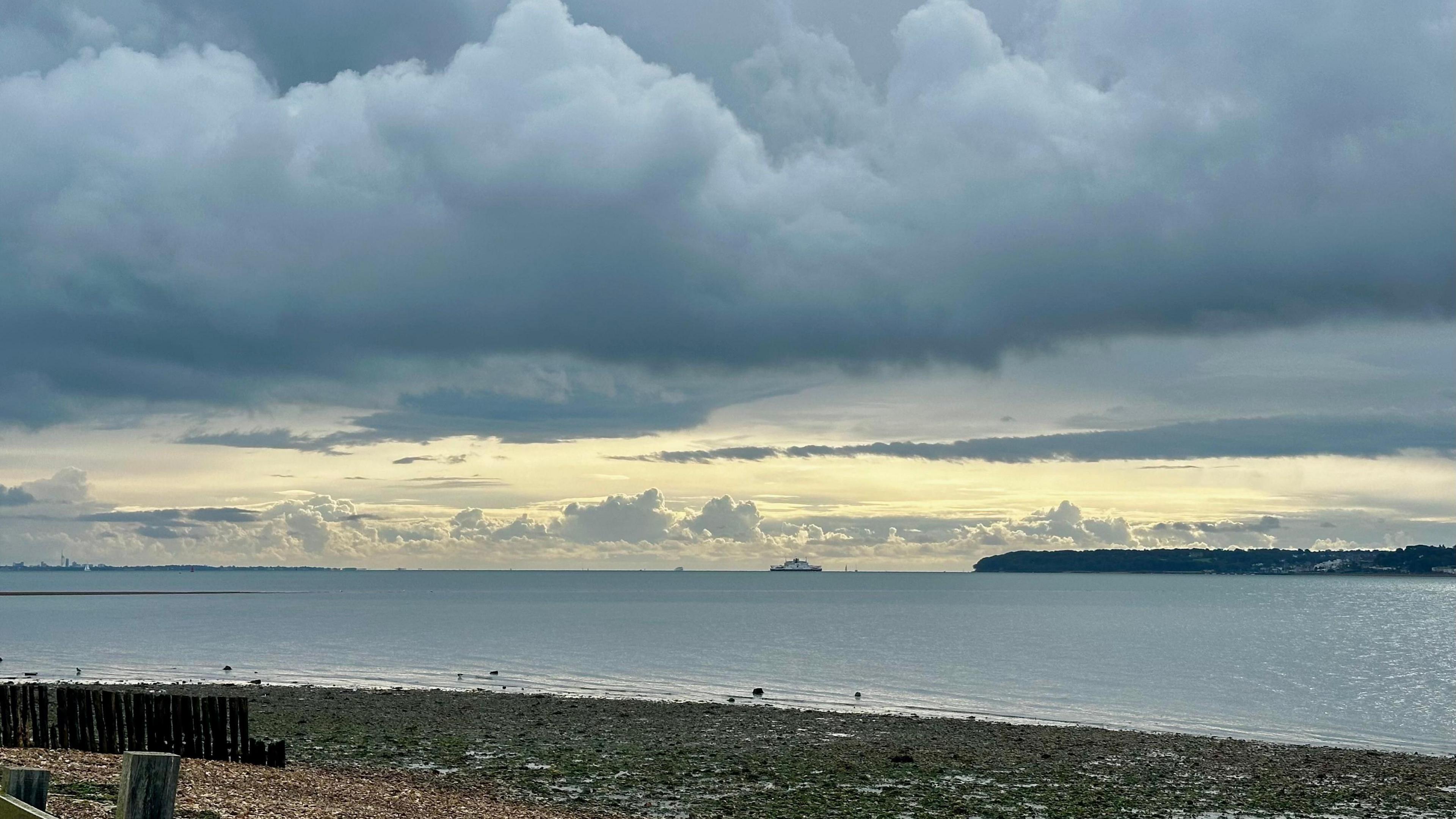 A beach scene but the sky is dark grey with ominous looking clouds. There is a line of lighter sky in the distance. A ferry can been seen on the water in the far distance