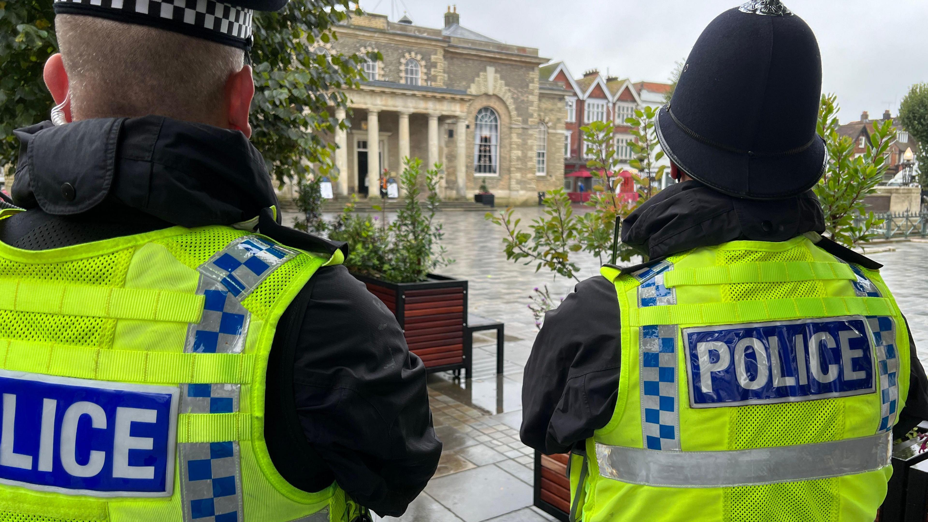 Two police officers overlook Salisbury market square. They are pictured from behind and are wearing high-vis jackets with "Police" in white letters on a blue panel on them, and traditional police helmets