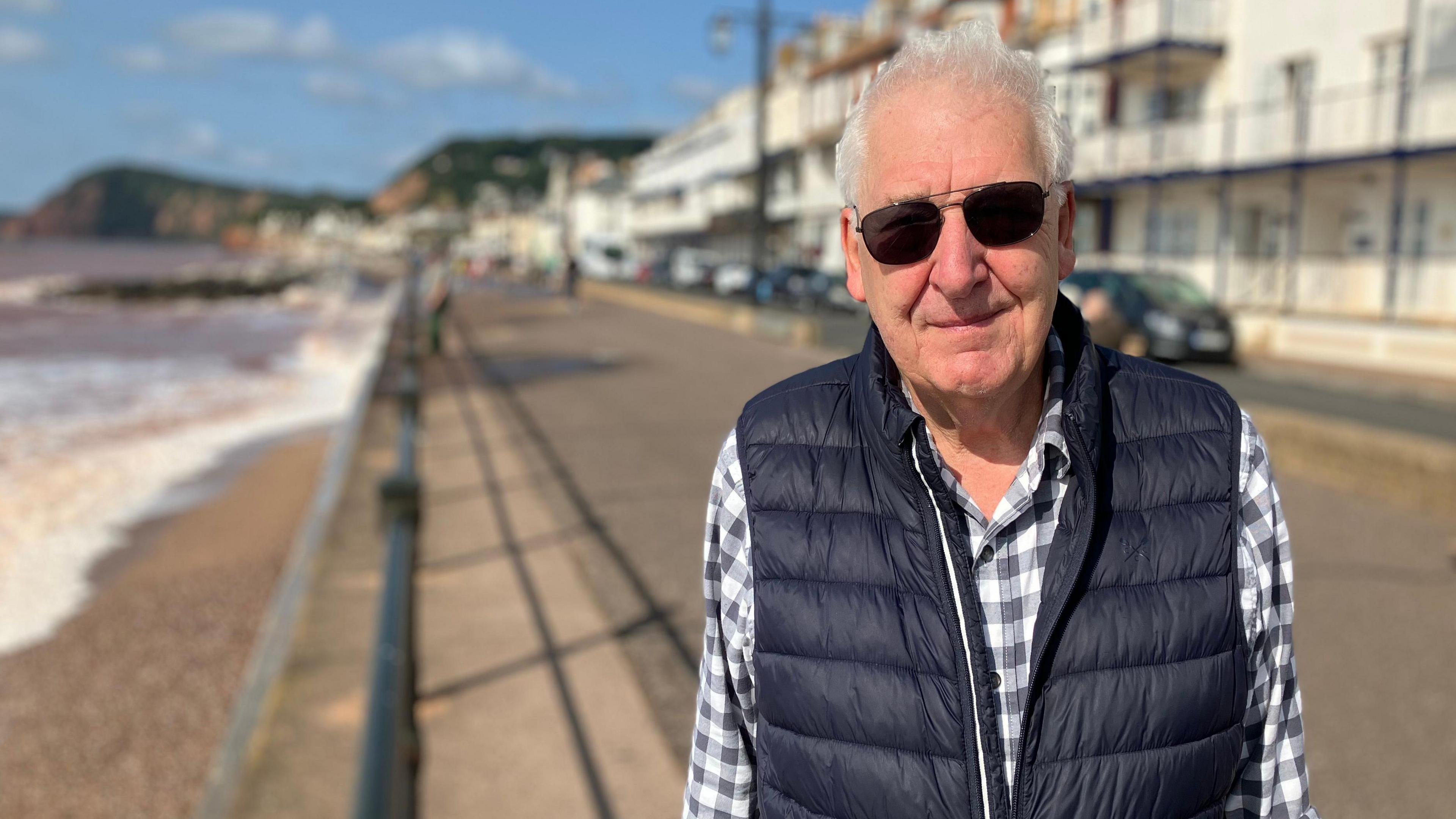 Geoff Jung, wearing a blue and white checked shirt, a gilet and sunglasses, standing on a sunny esplanade in the coastal town of Sidmouth, with the promenade behind him and waves breaking on the beach