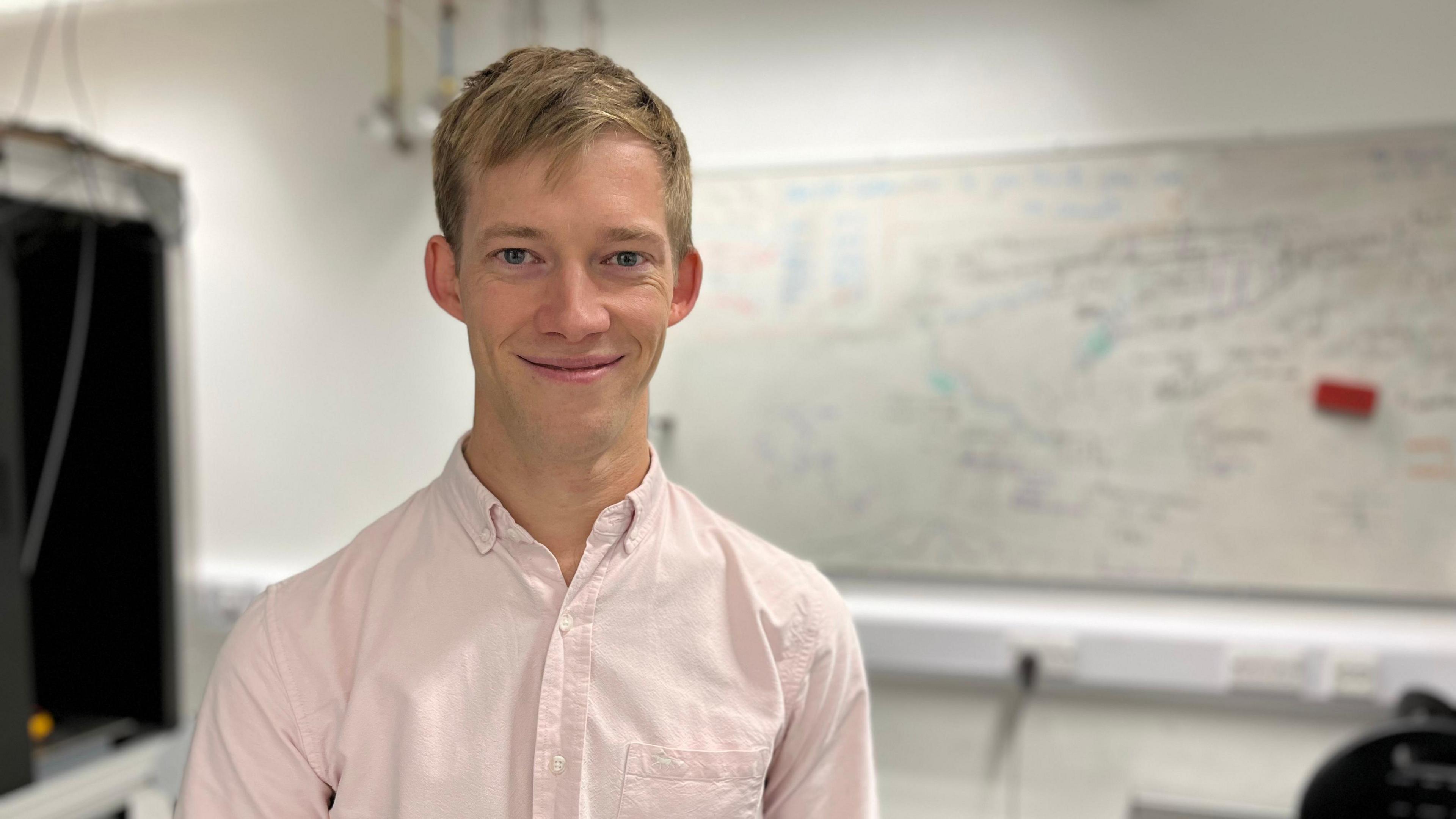 A blonde man in a pale pink shirt standing in front of a whiteboard with writing on it