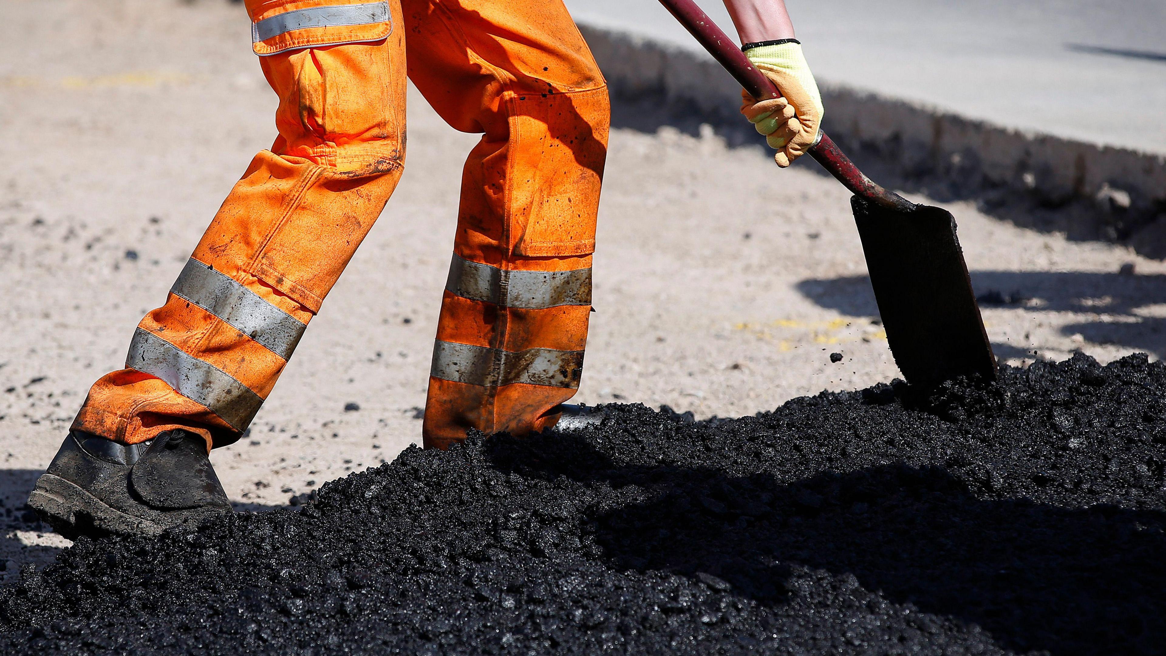 The bottom half of a worker wearing orange hi-vis trousers, laying tarmac on a road.