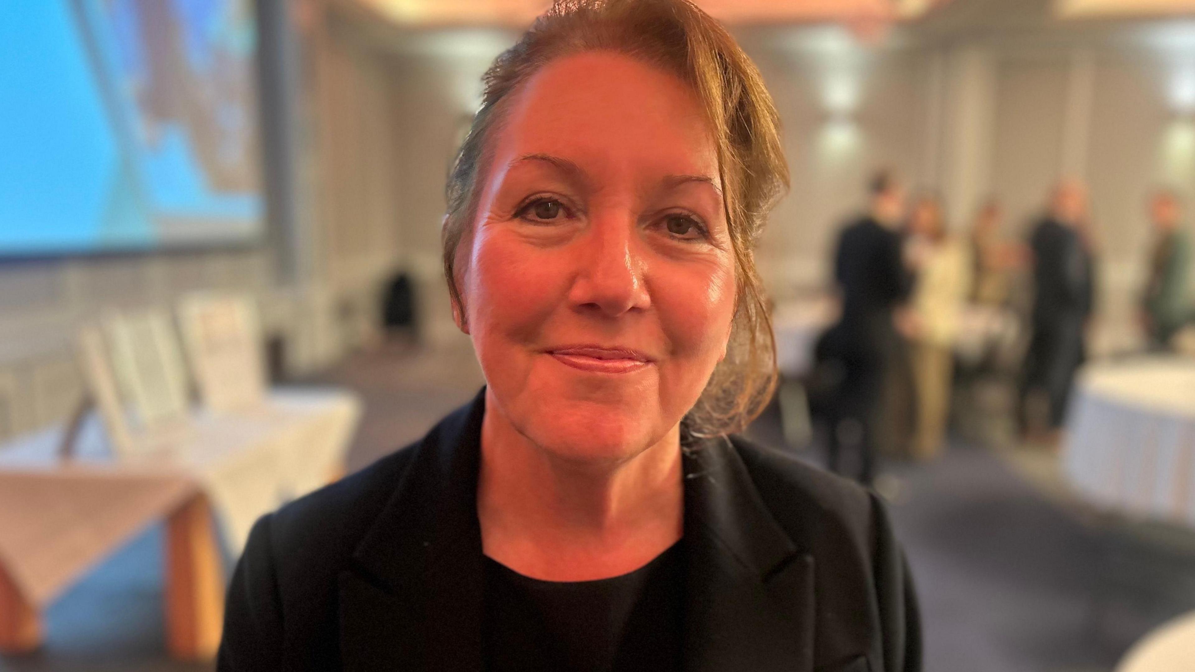 Jan Cox smiles at the conference, her brown hair is tied back off her face. She wears a black jacket and top.