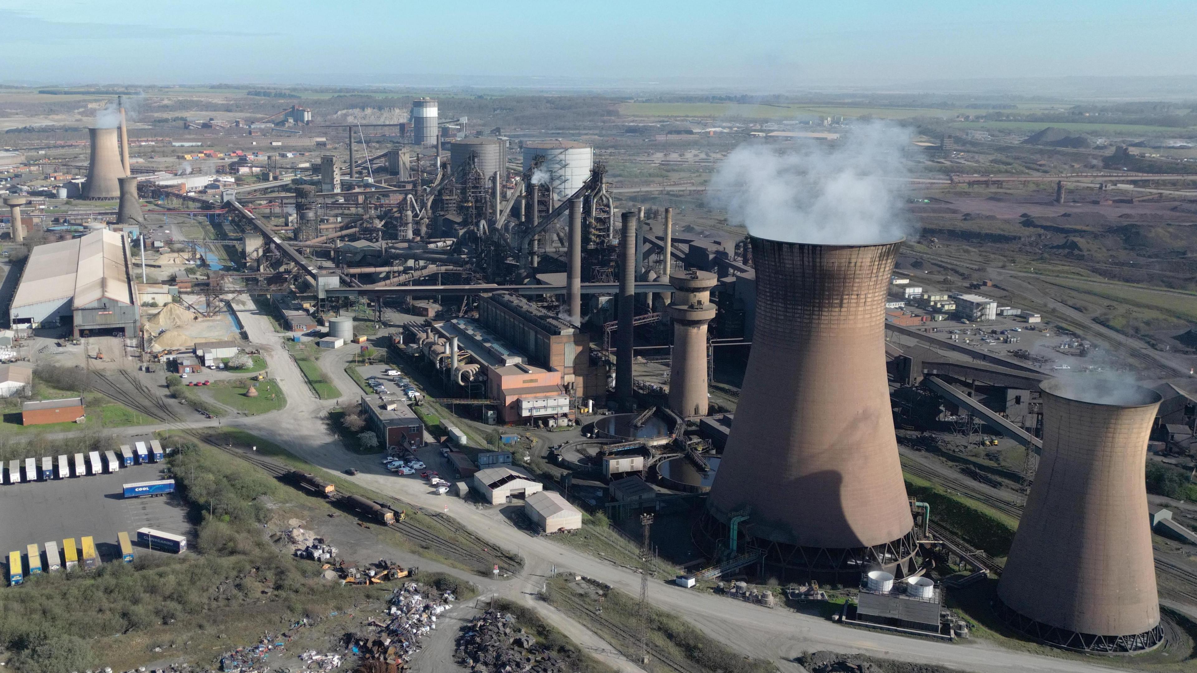 An aerial view of the British Steel plant in Scunthorpe. Two large brown blast furnaces eject smoke beside a sprawling plant of brown chimneys and pipes, set  against rolling fields in the background.
