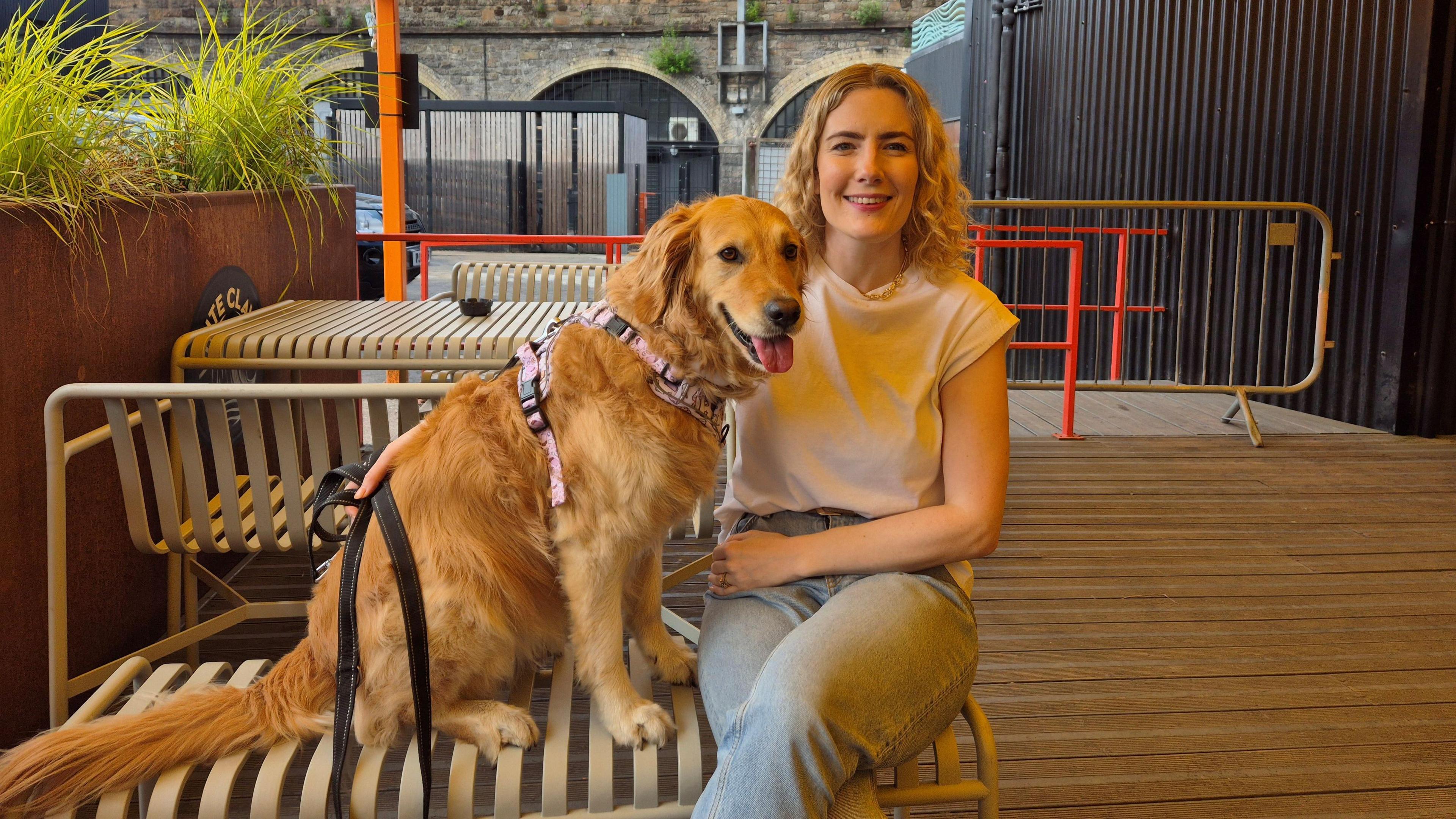 Jenn Nimmo-Smith - a woman with long blonde hair - sits on a bench in a bar's outdoor area alongside her dog Scout, a large golden retriever