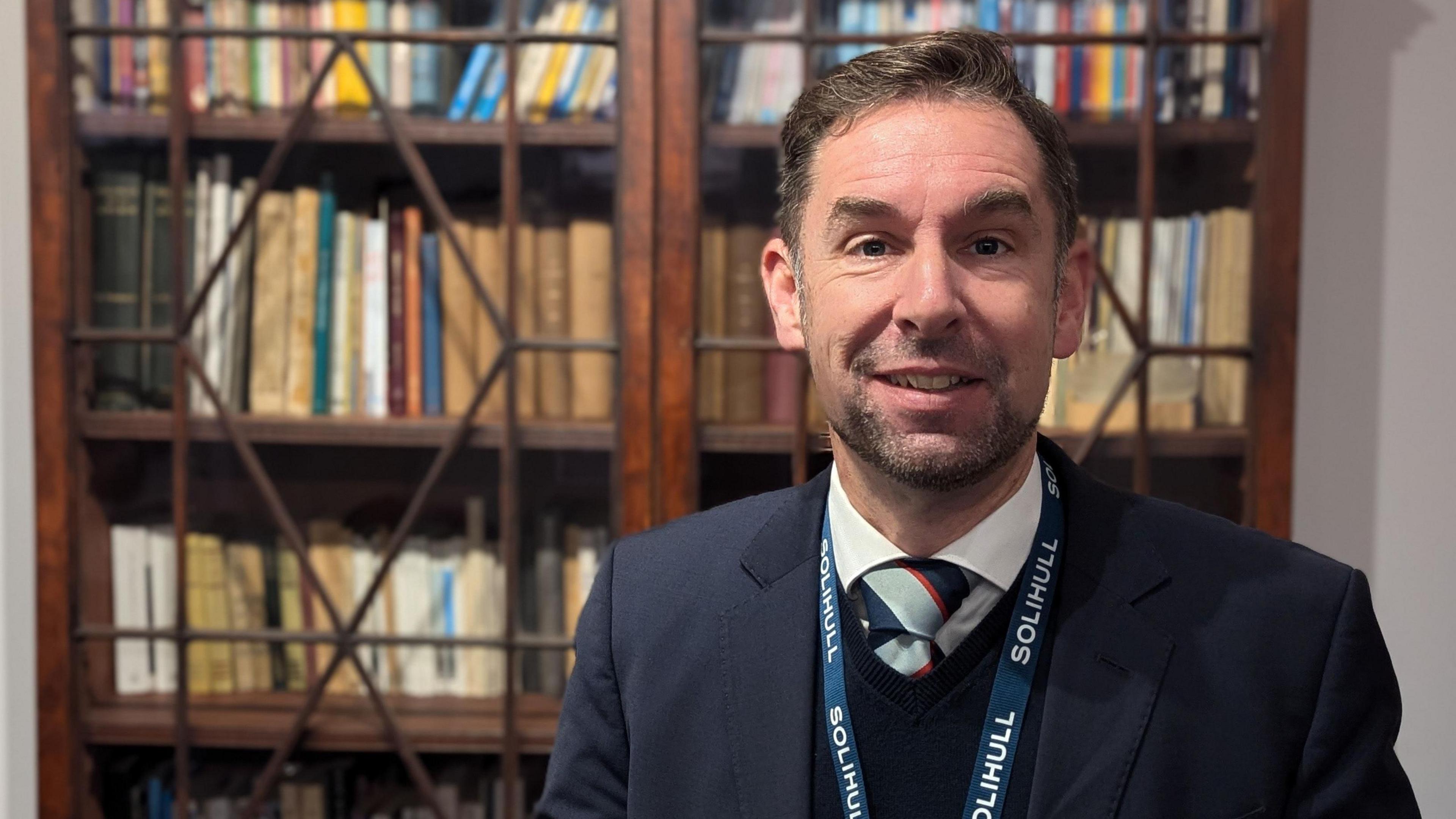 A man stands in front of a bookcase. He has brown hair and a beard, and he is wearing a dark blue suit. He has a lanyard around his neck that says "SOLIHULL" in capital letters on the neck ribbon.