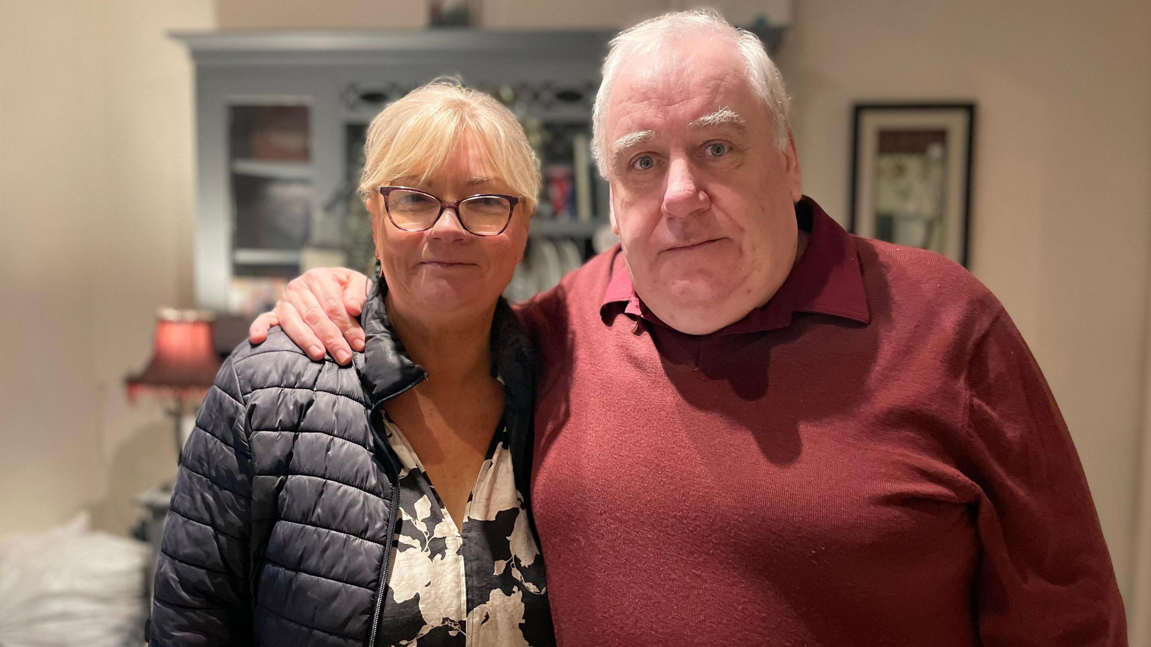 A woman with blonde hair wearing a blue coat and floral white and black shirt. She is wearing black-rimmed glasses and is stood next to a man wearing a dark red jumper and polo shirt who has his arm around her and stood inside a room in their home. He has white hair and eyebrows.