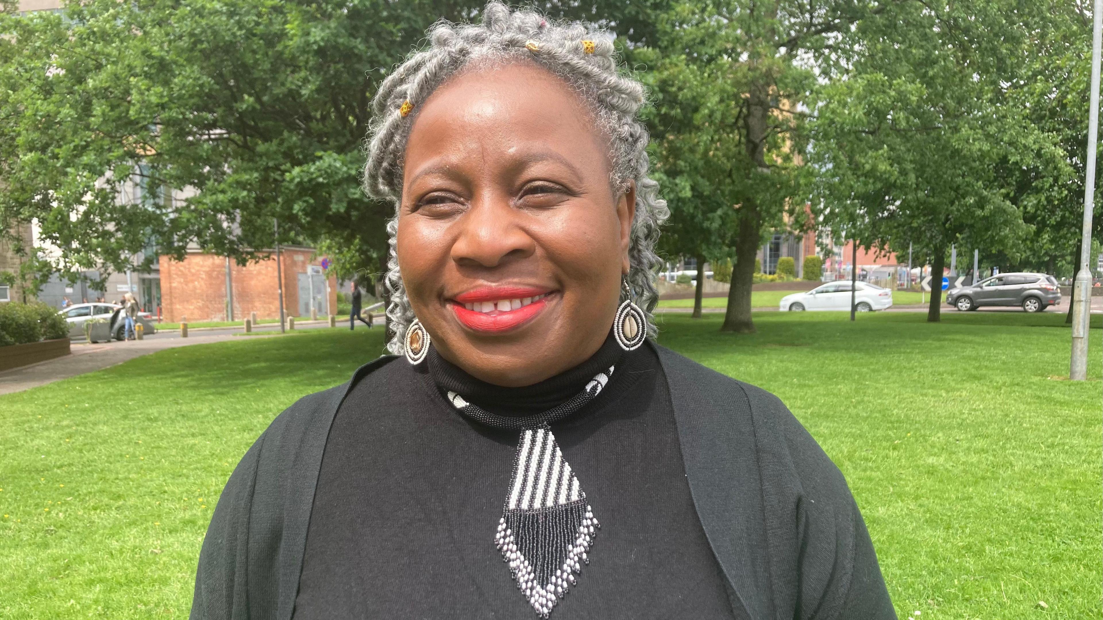 Beverly Simpson is smiling into the camera with some trees and grass in the background. She is wearing a black top and large earrings 