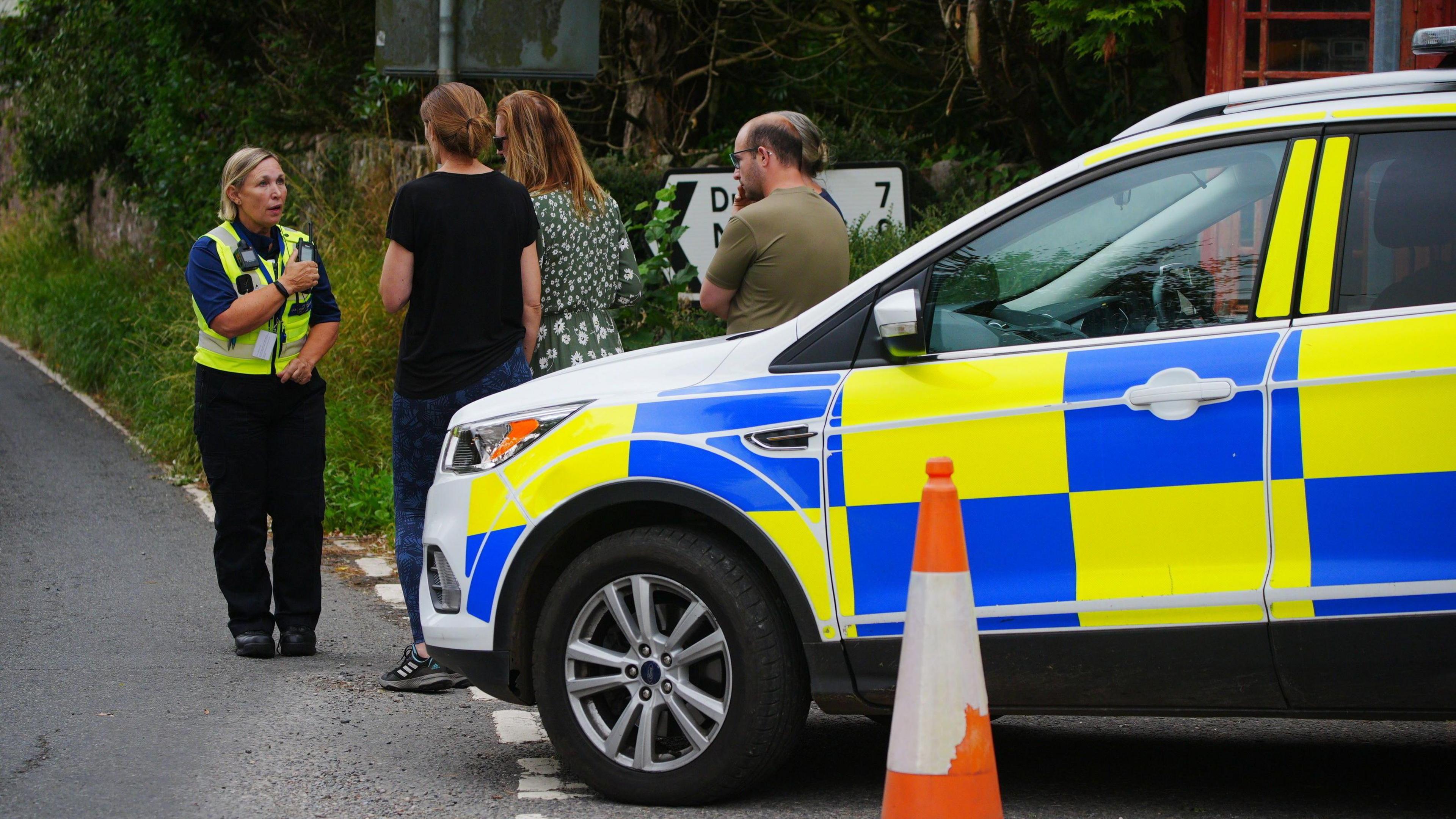 A female police officer in uniform talks to local residents standing next to a police car in Wheddon Cross in Somerset after a coach crash