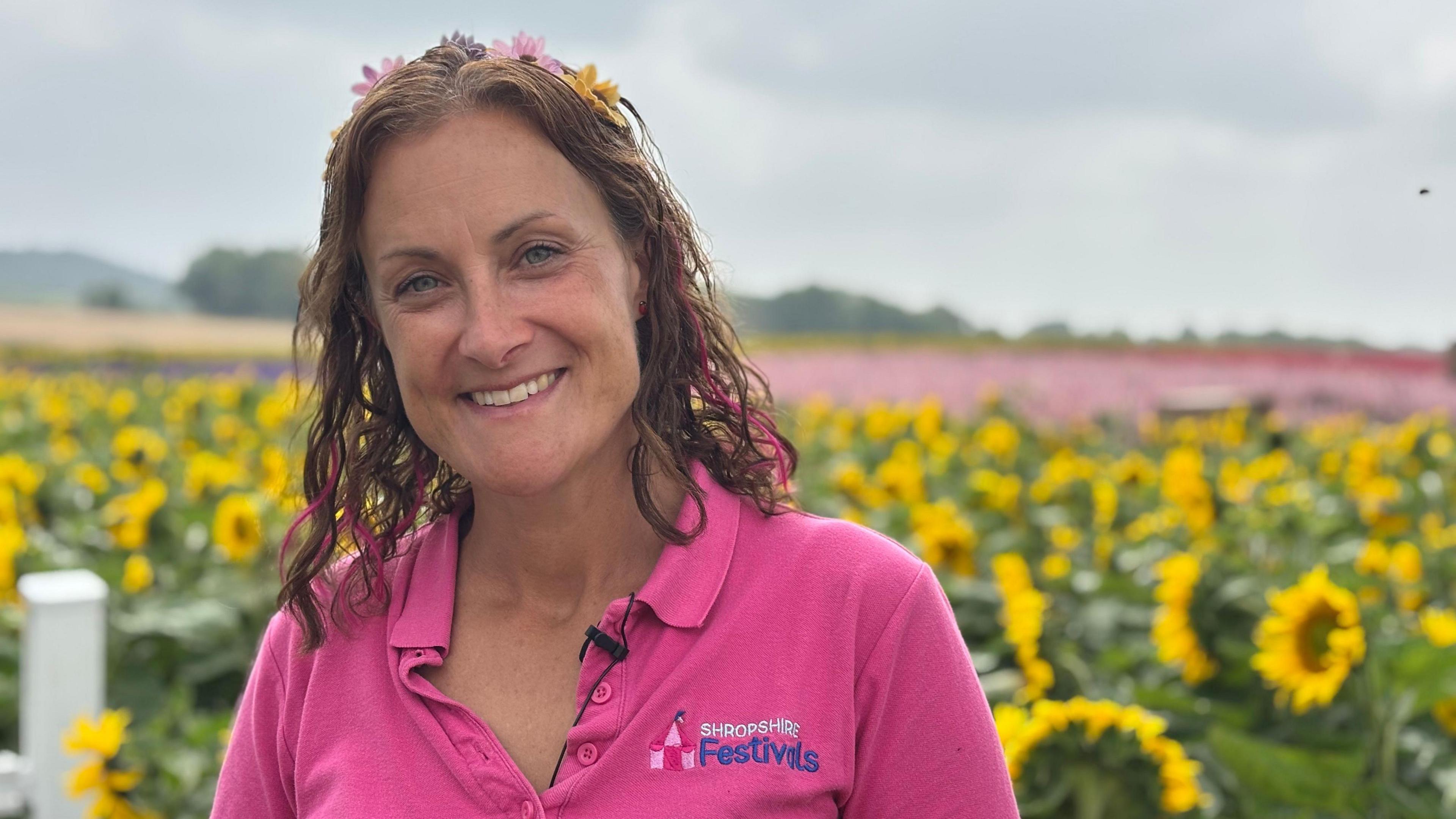 A woman with brown hair with pink streaks. and a yellow and pink flower crown in her hair. She is wearing a pink t-shirt with the Shropshire Festivals logo on it. In the background is a sea of sunflowers, and beyond that, pink flowers