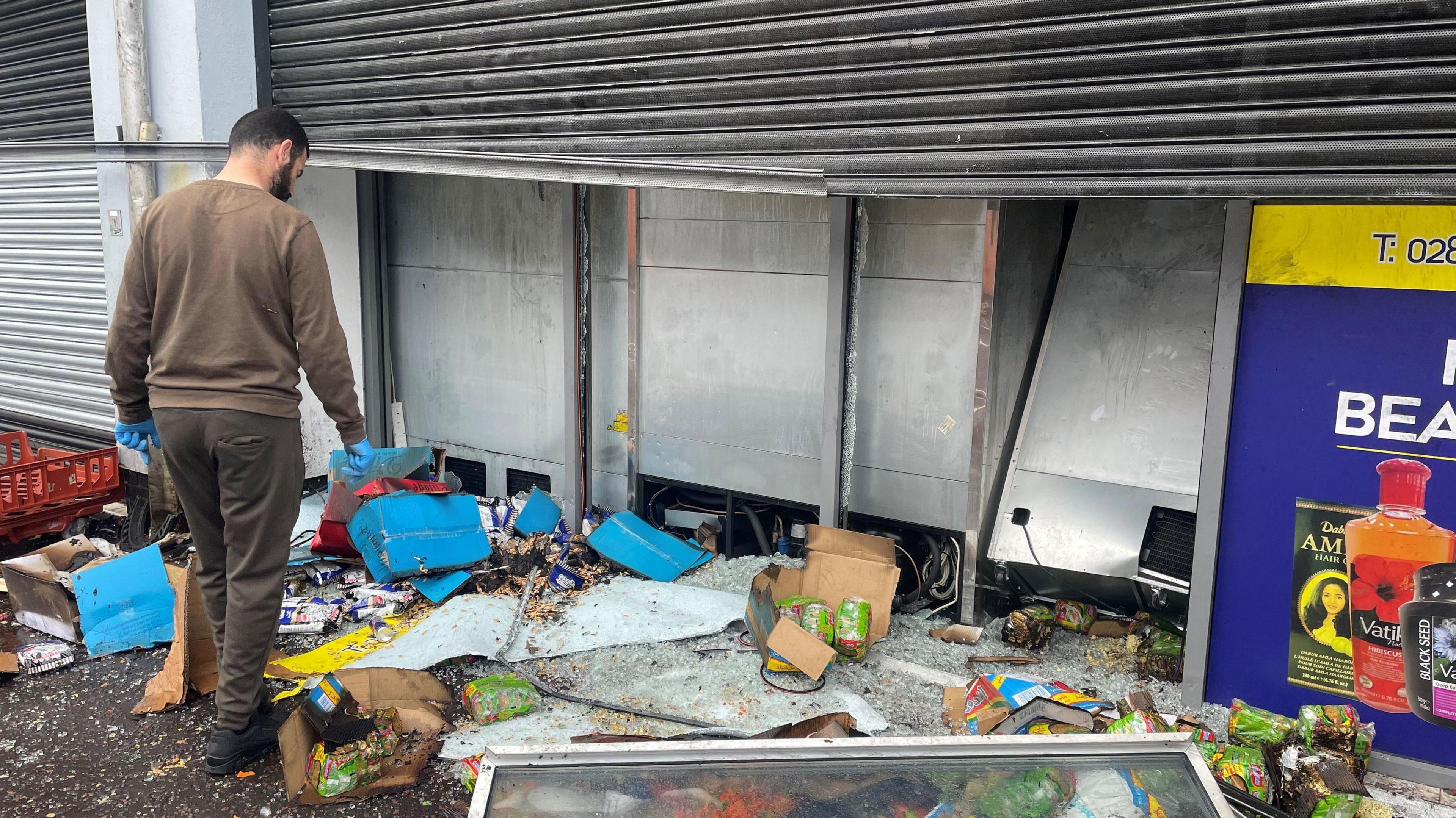 Damaged supermarket on Donegall Road, groceries and glass panel strewn across pavement, man in brown jumper inspecting the scene