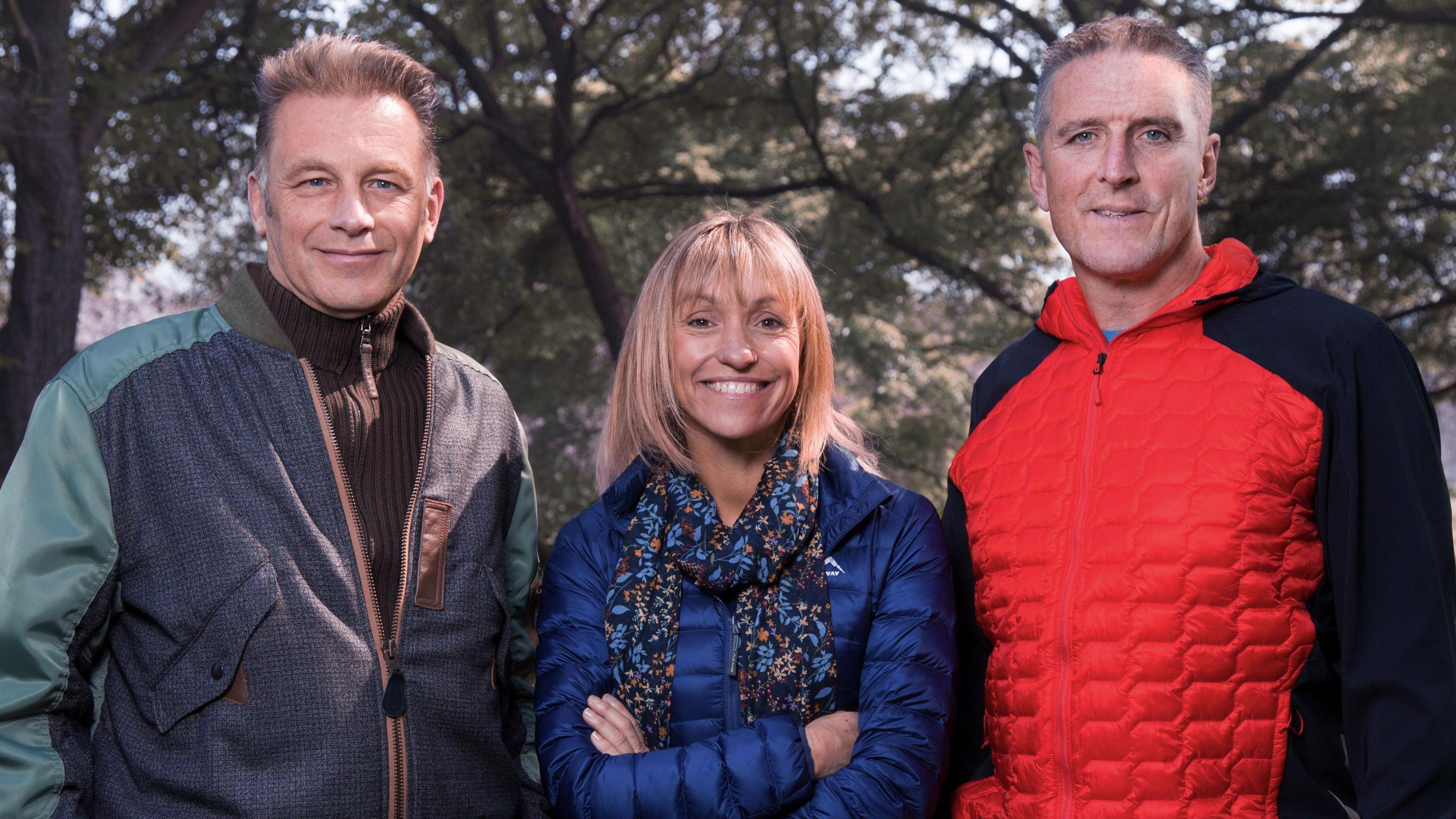 Chris Packham, Michaela Strachan and Iolo Williams stand in a row in front of trees and smile at the camera. On the right, Chris Packham, who has short, light coloured hair, is wearing a brown ribbed jumper and a patchwork zip-up jacket. In the middle, Michaela has blonde, medium-length hair with a full fringe and is wearing a blue puffed jacket and a blue and orange patterned scarf. She has her arms crossed across her chest. On the right, Iolo has short grey hair and is wearing a red and black padded zip up raincoat.