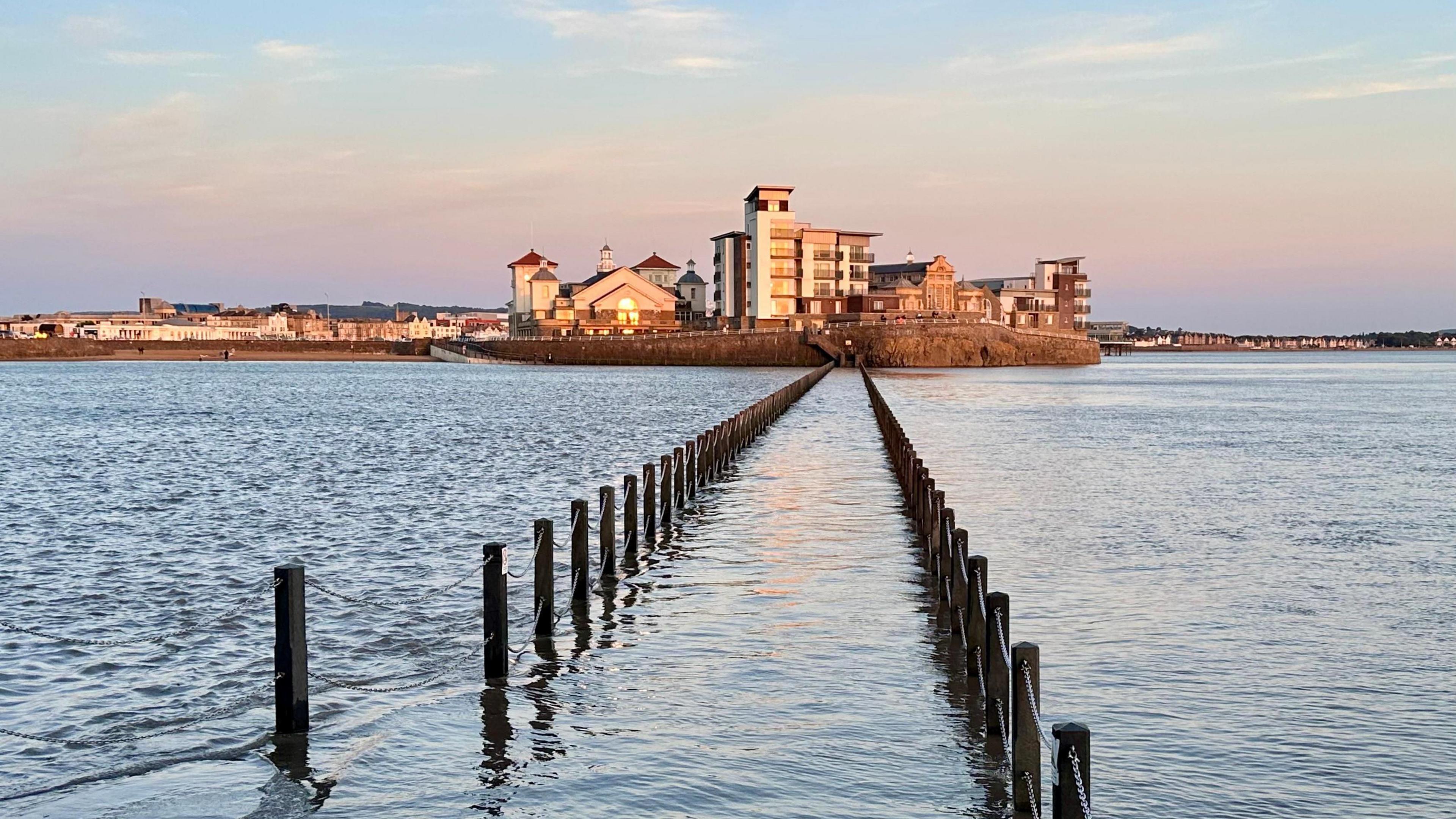 A view over the sea during sundown. There are pillars leading to one side of the beach, where the sun is hitting the windows of houses, making them light up.