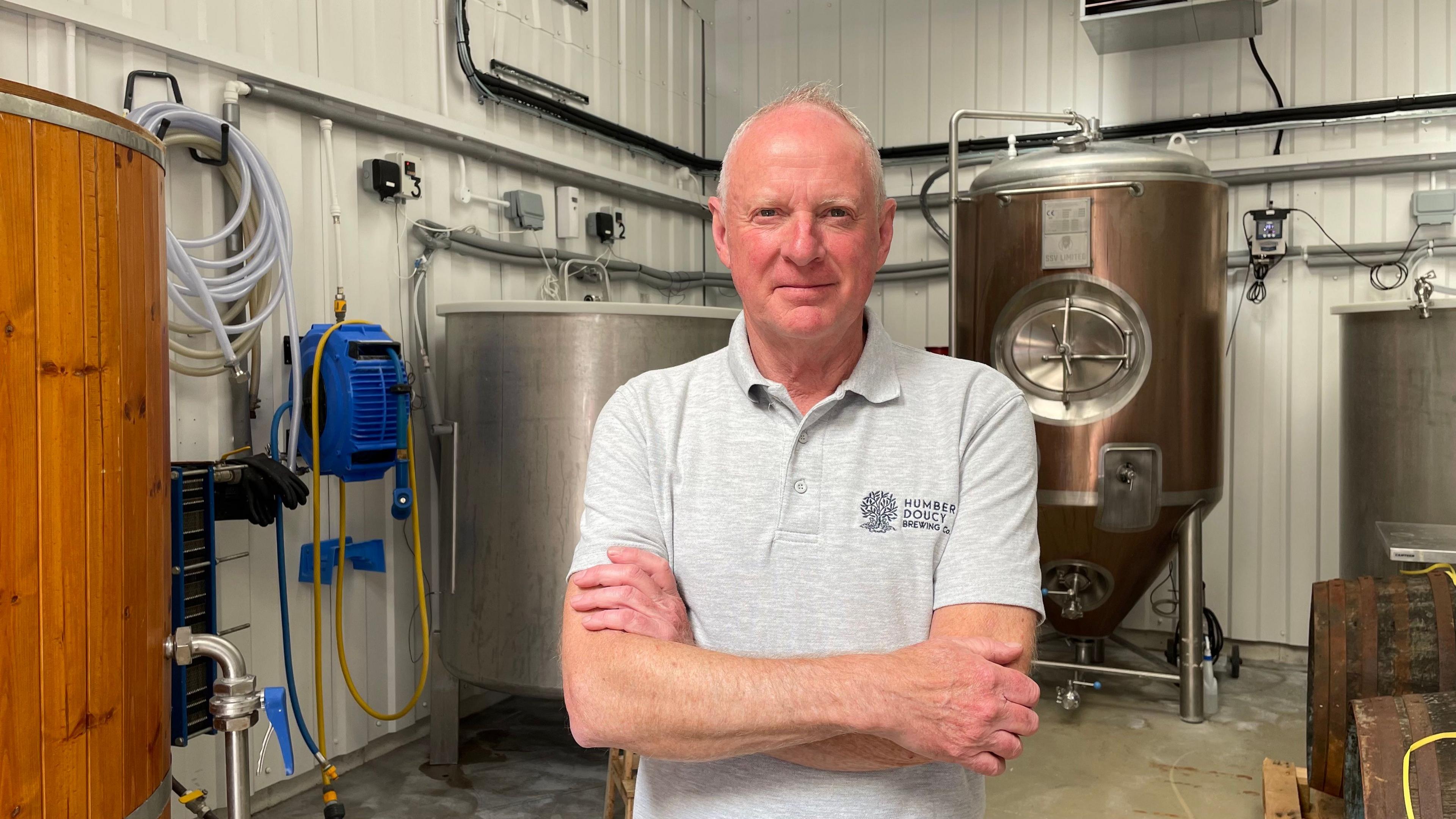 Alan Ridealgh, standing in his brewery's brewhouse, looking straight to camera. He is wearing a grey T-shirt, with Humber Doucy written on the chest. His arms are folded and he has short, grey hair. A large wooden container is seen on the left of frame, while in the background are three metal containers, which are used in the brewing process. The walls are white corrugated metal.