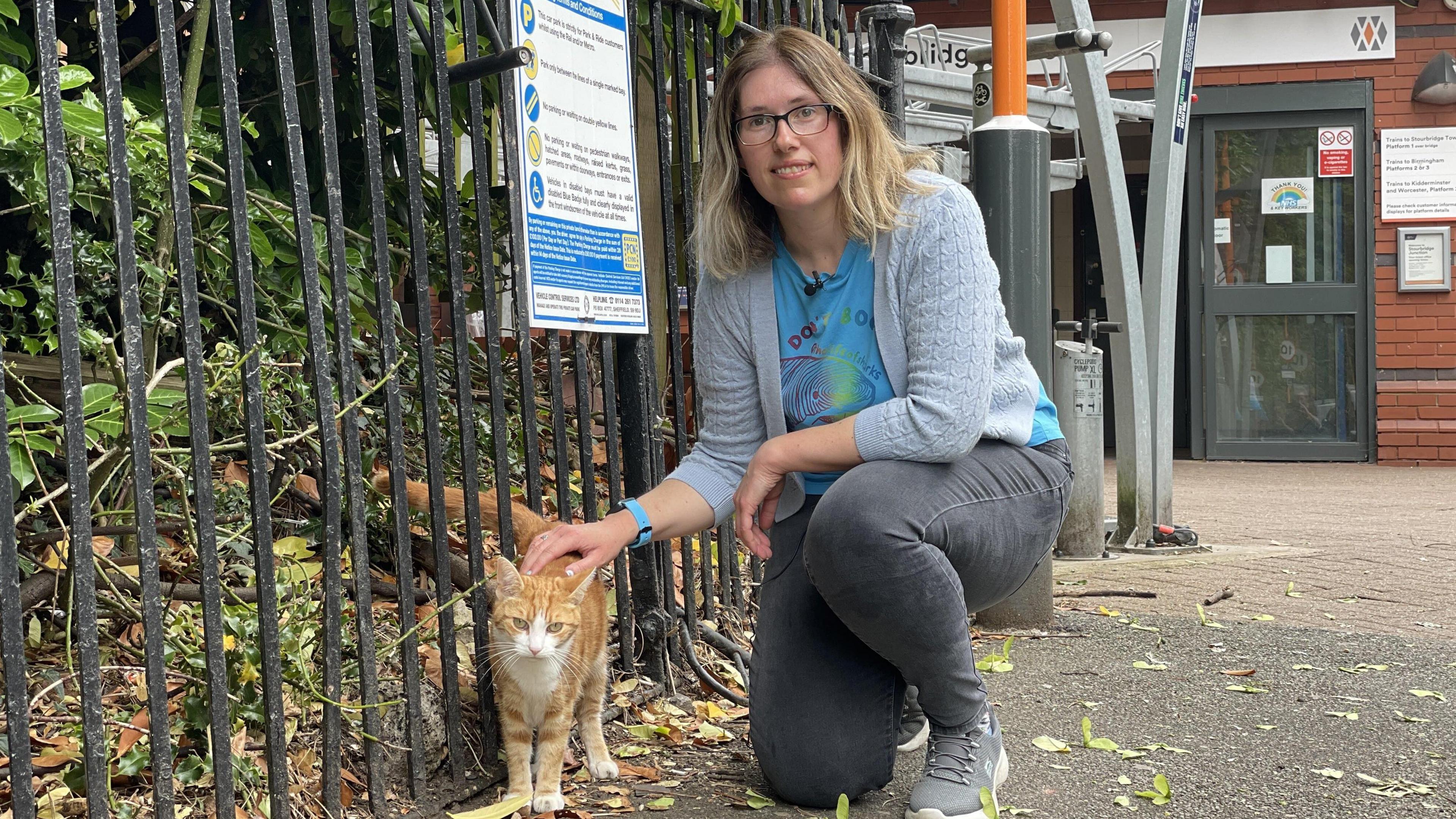 A woman in a grey cardigan, blue t-shirt and grey jeans crouches next to a ginger and white cat, stroking its back. They are outside a train station, next to a metal fence