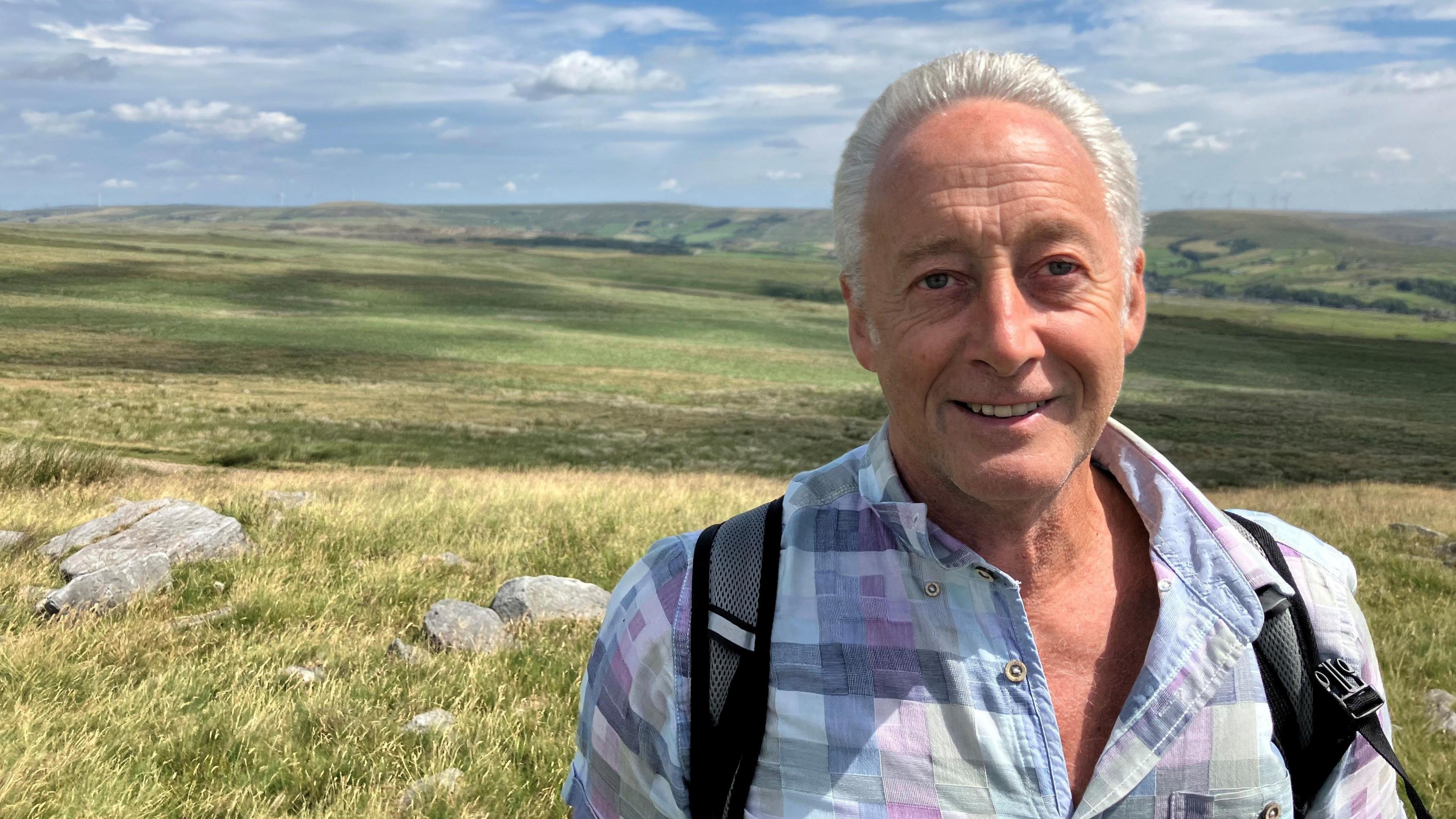 Photograph of Stuart Davies from the Rooley Moor Neighbourhood Forum. The picture is taken on the Cotton Famine Road above Rochdale. He has swept-back grey hair and is wearing an open-necked checked shirt in shades of blue, green and purple.