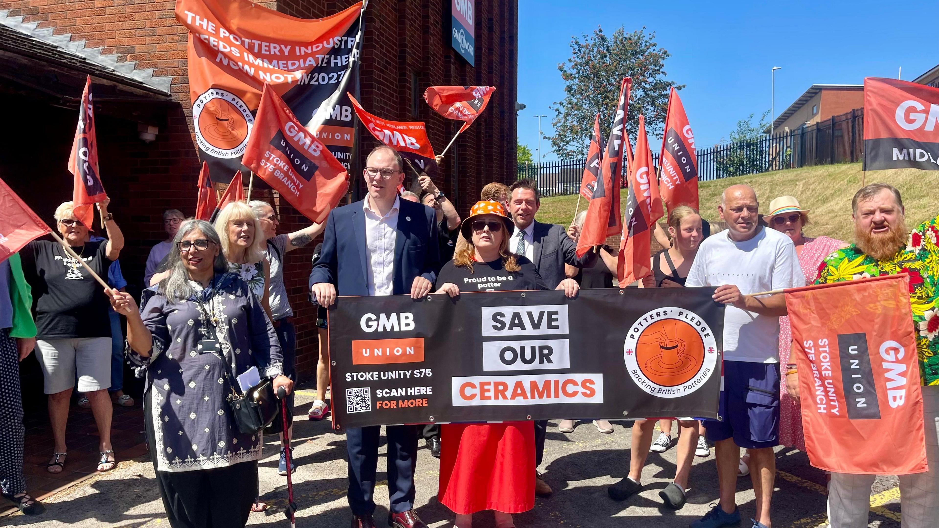 A group of people holding banners and flags. One of the flags reads 'Save Our Ceramics'. The flags bear the GMB union's logo on an orange background.