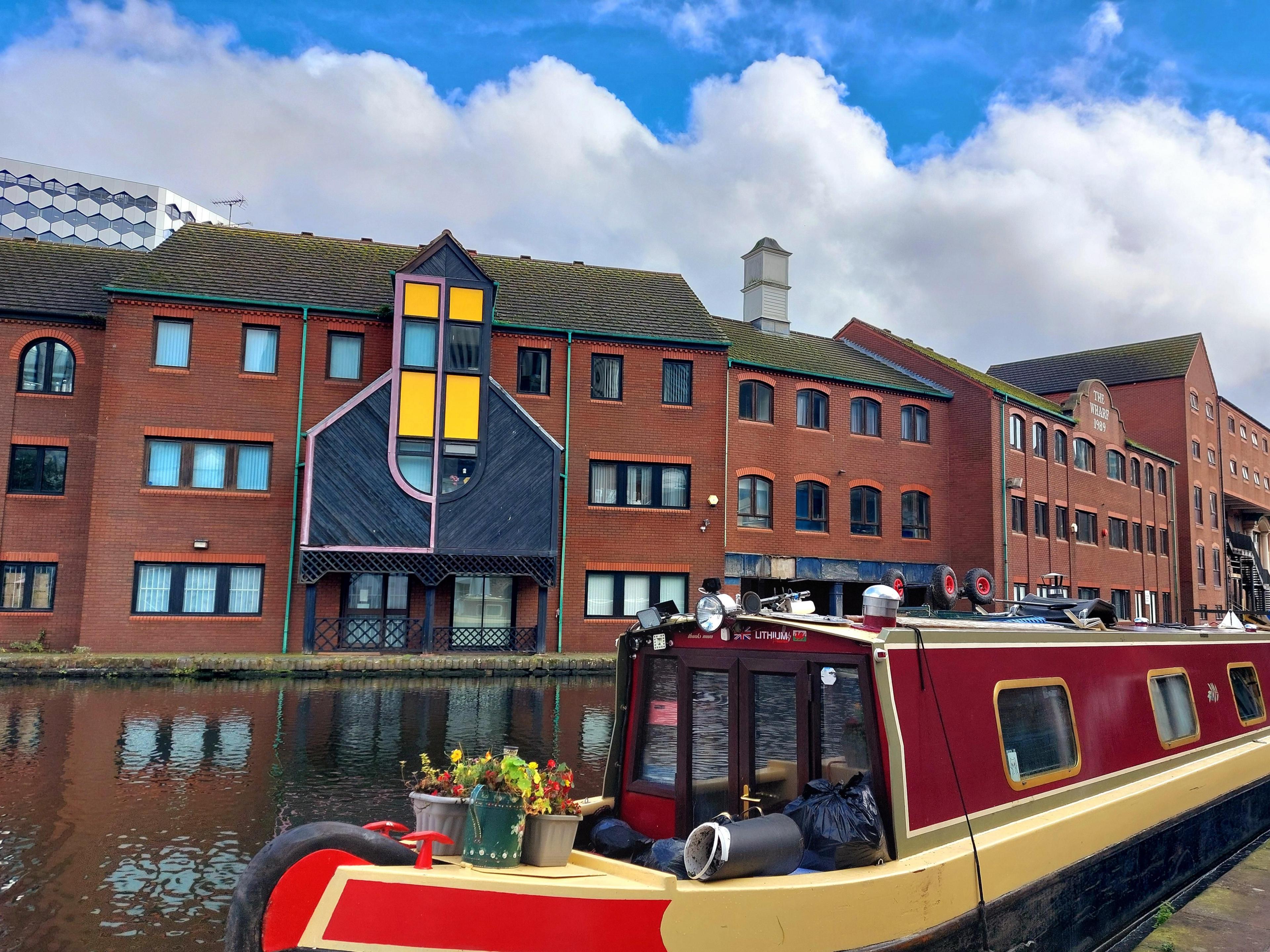 A red and yellow canal boat with buckets of flowers on the prow. Behind, modern three-storey flats built in red brick with colourful wood detail.