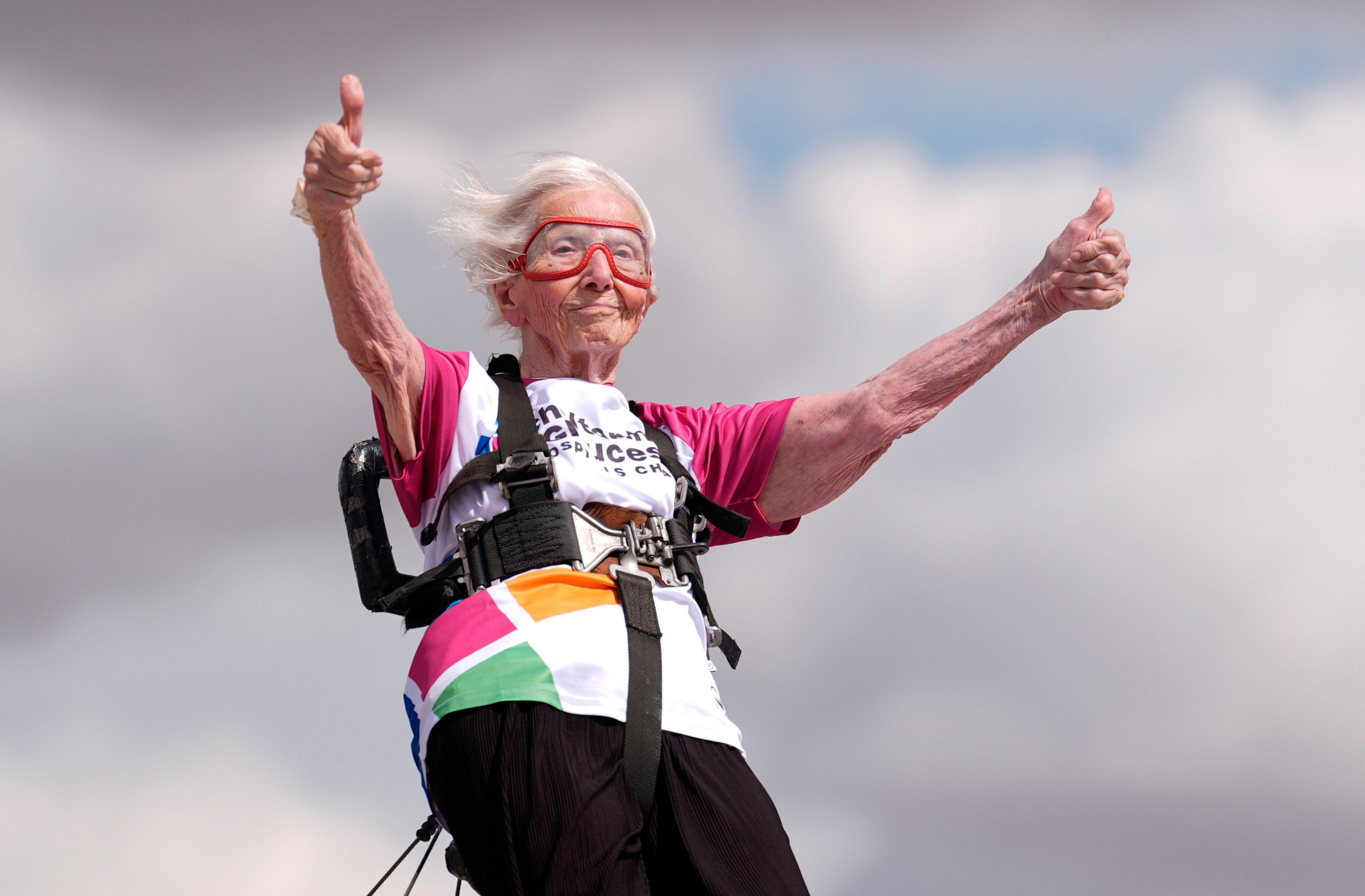 97-year-old Betty Bromage wing walks on a plane. She wears goggles and gives two thumbs up.