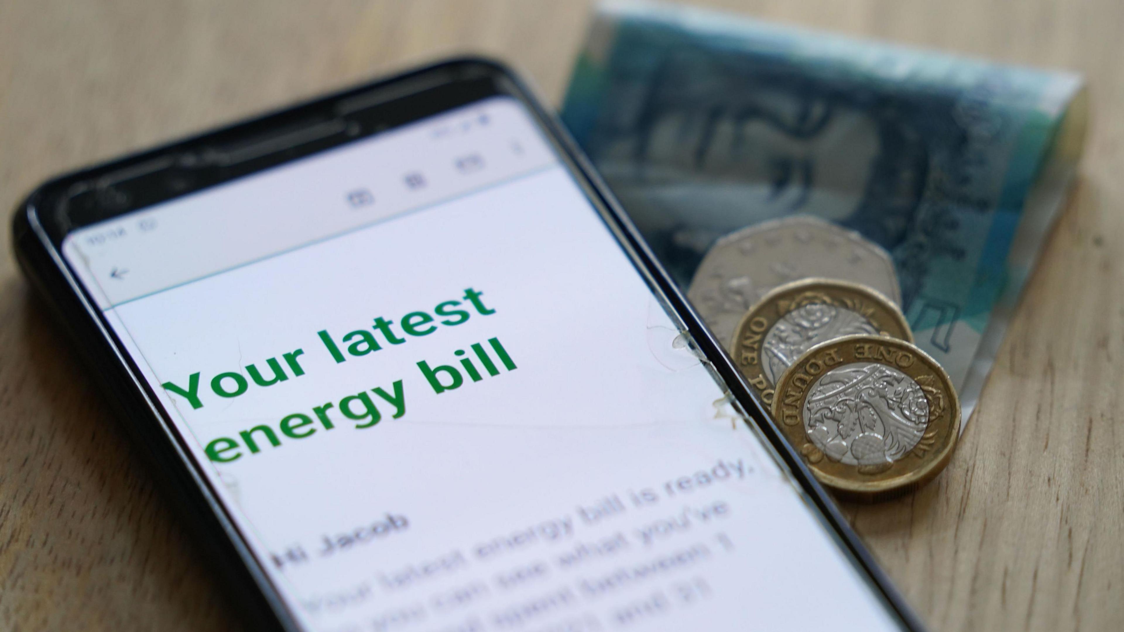 A letter which is being read on a mobile phone. The phone is laid on a wooden table with two one pound coins, a 50p and a £5 note. On the phone it says "Your latest energy bill".